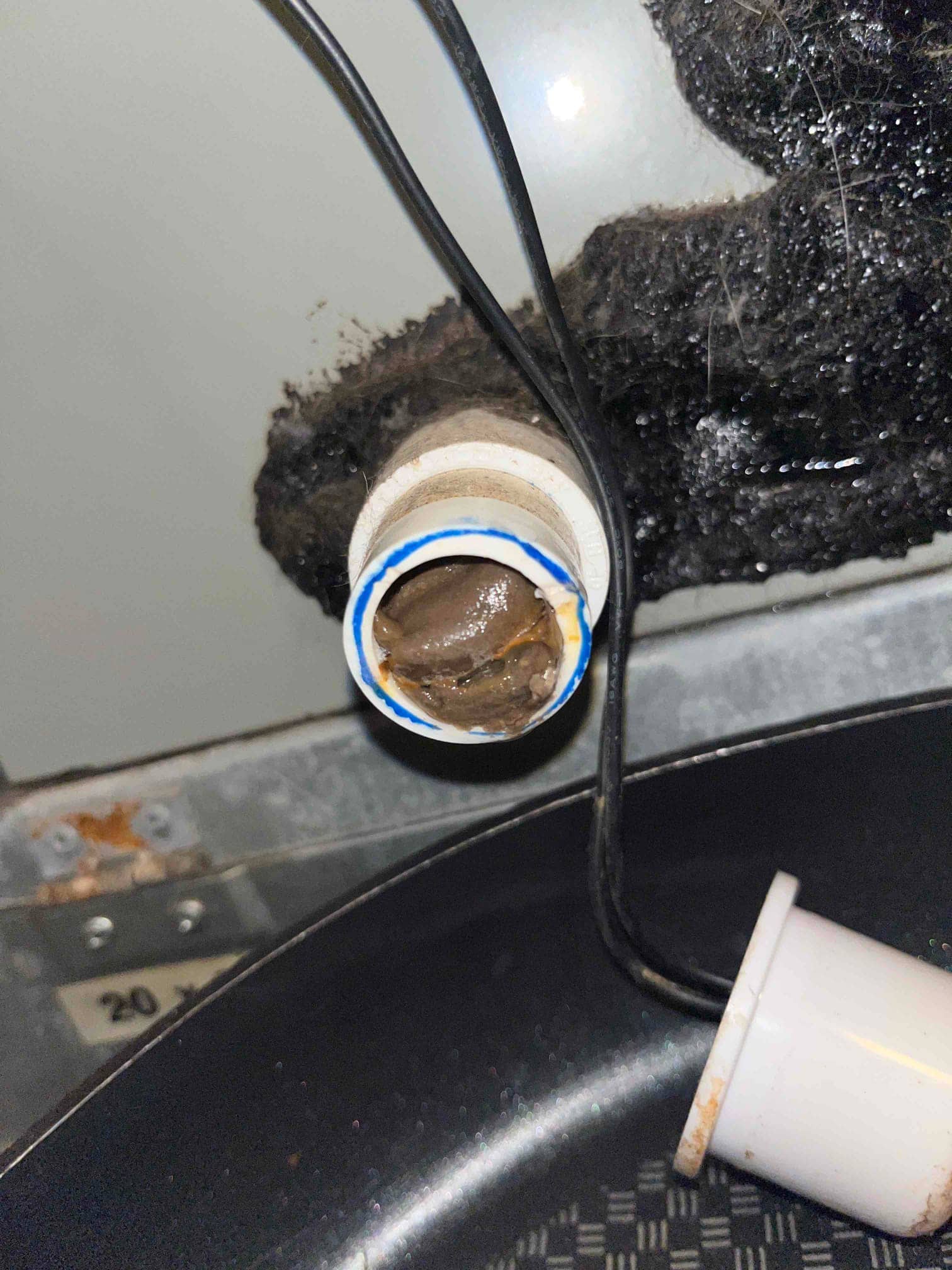 Clogged condensate drain fitting with brown sludge buildup, adjacent to insulated drain line and drip pan inside the air handler.
