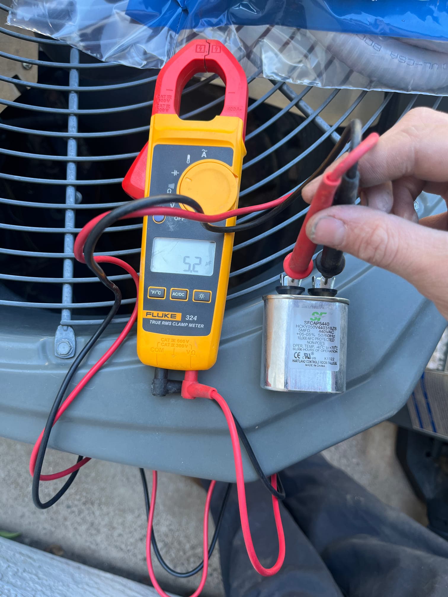 Digital multimeter measures 5.2 µF on the removed capacitor using test leads next to the condenser unit.