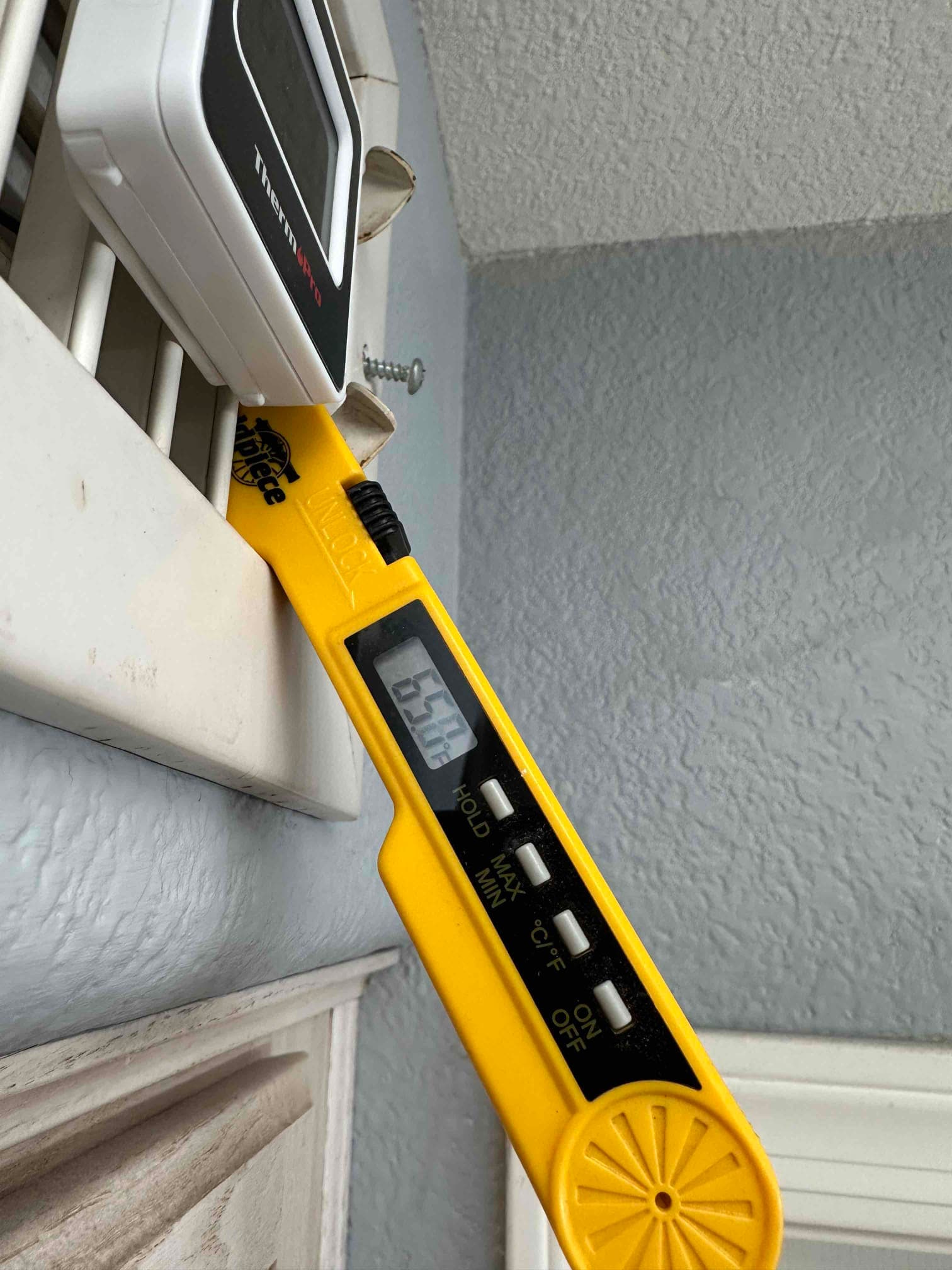 Digital thermometer measures supply air temperature at a wall register, with the display reading 65.7 degrees Fahrenheit.