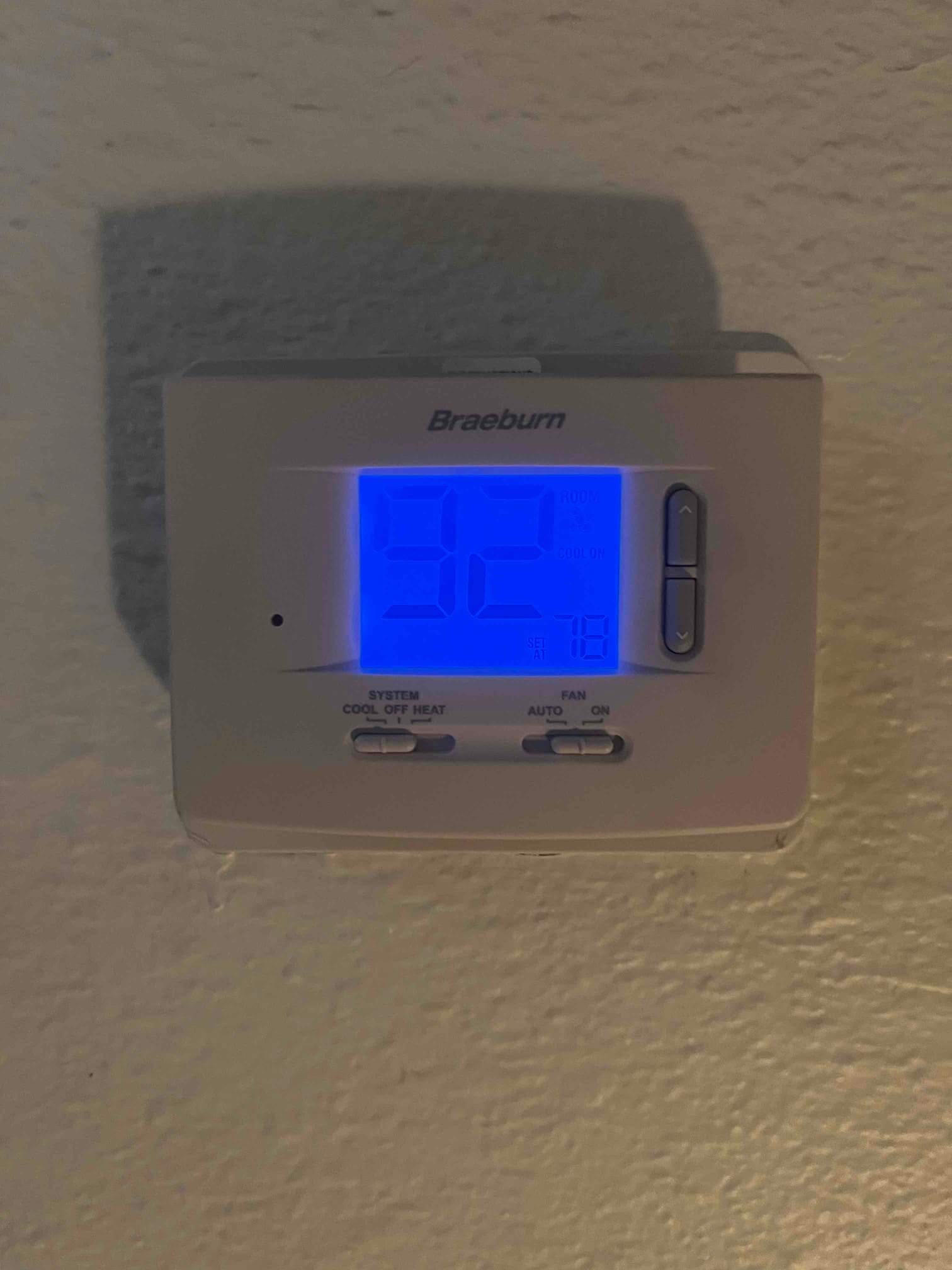 Digital thermostat on a wall displays the current setpoint and room temperature with system and fan mode buttons.