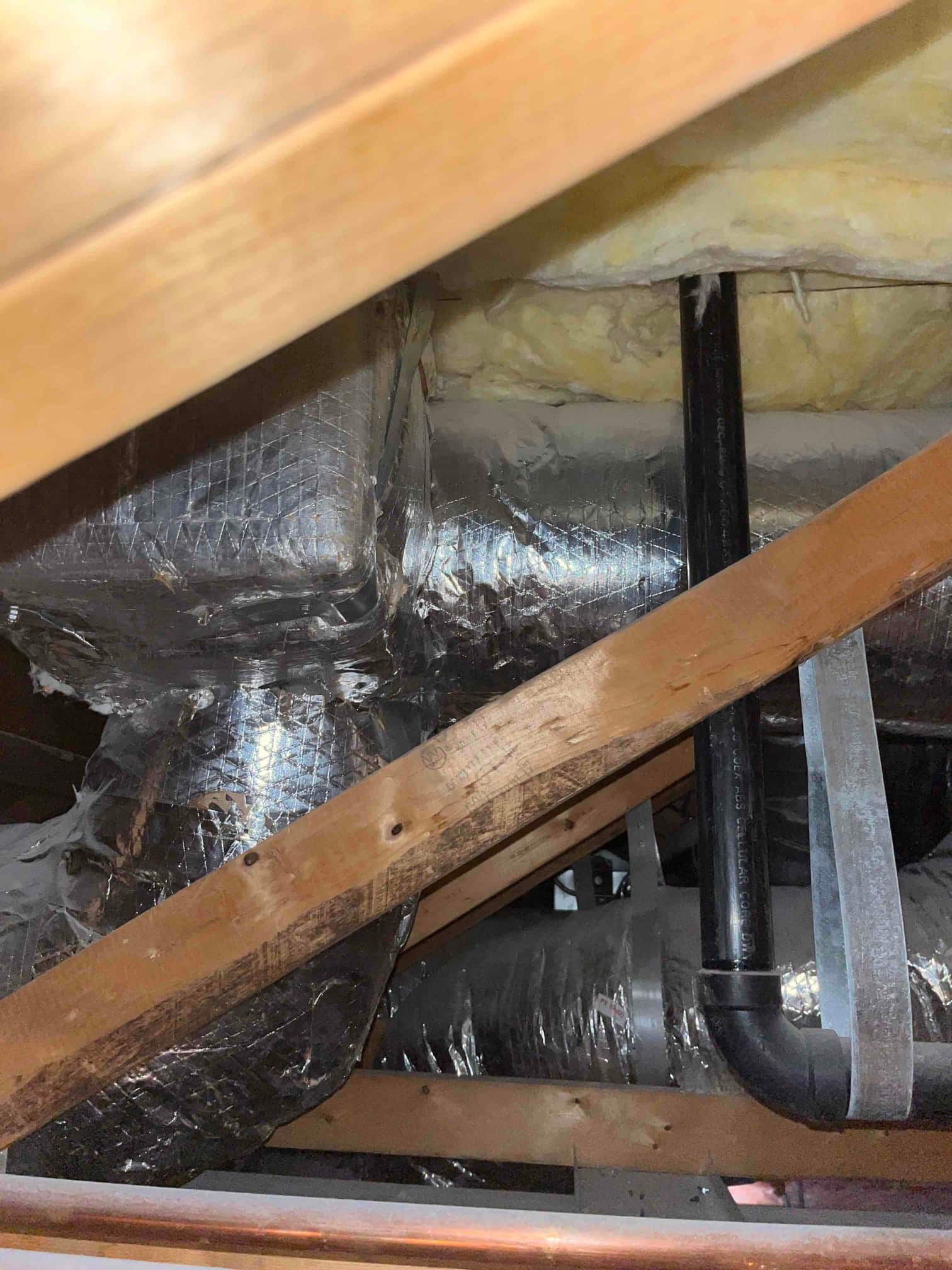 Insulated ductwork and PVC condensate drain piping run through an attic space with visible wooden framing and insulation.