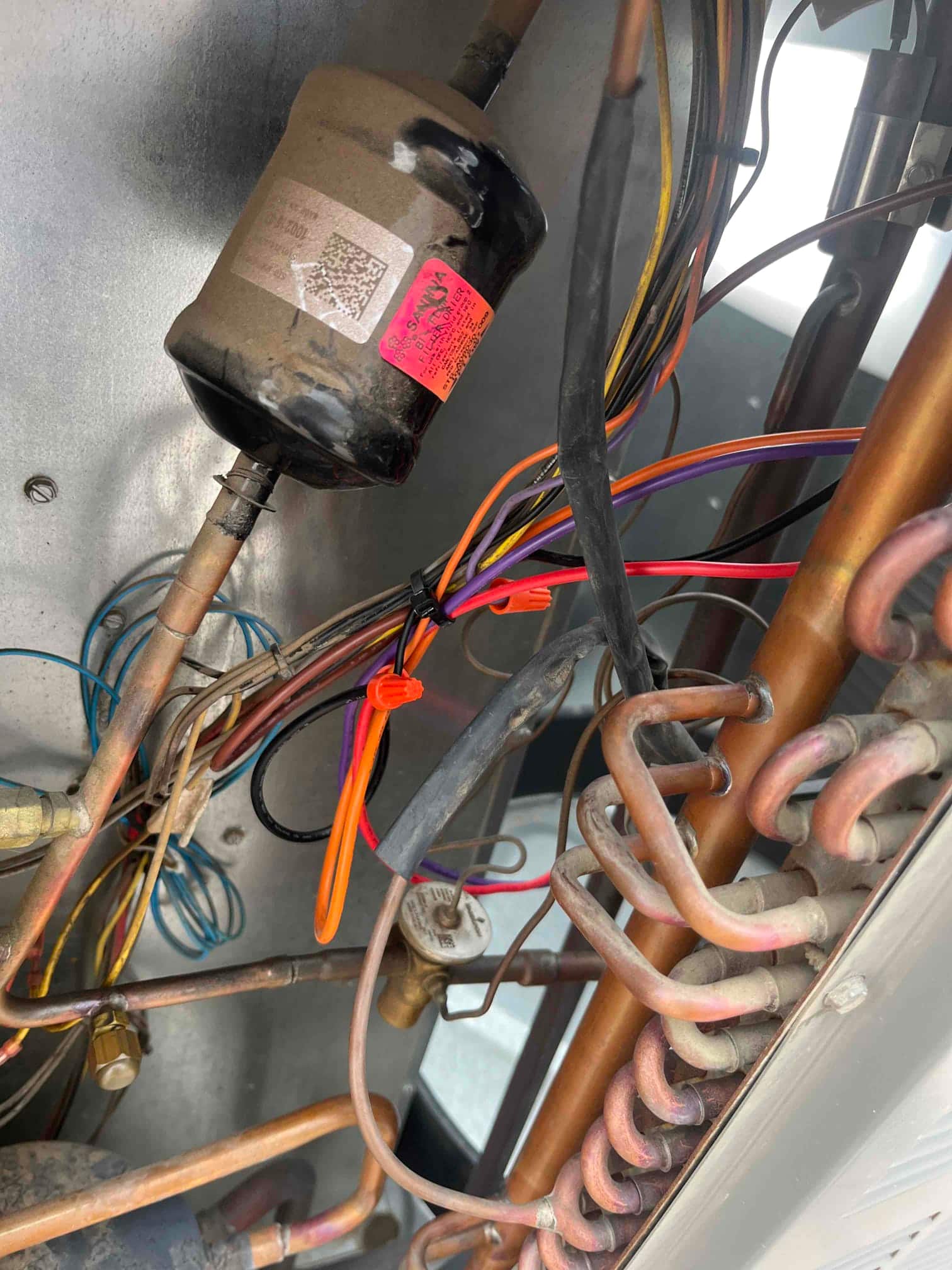 Interior of an outdoor condenser cabinet showing refrigerant lines, filter-drier, wiring harness with zip ties, and copper coil tubing.