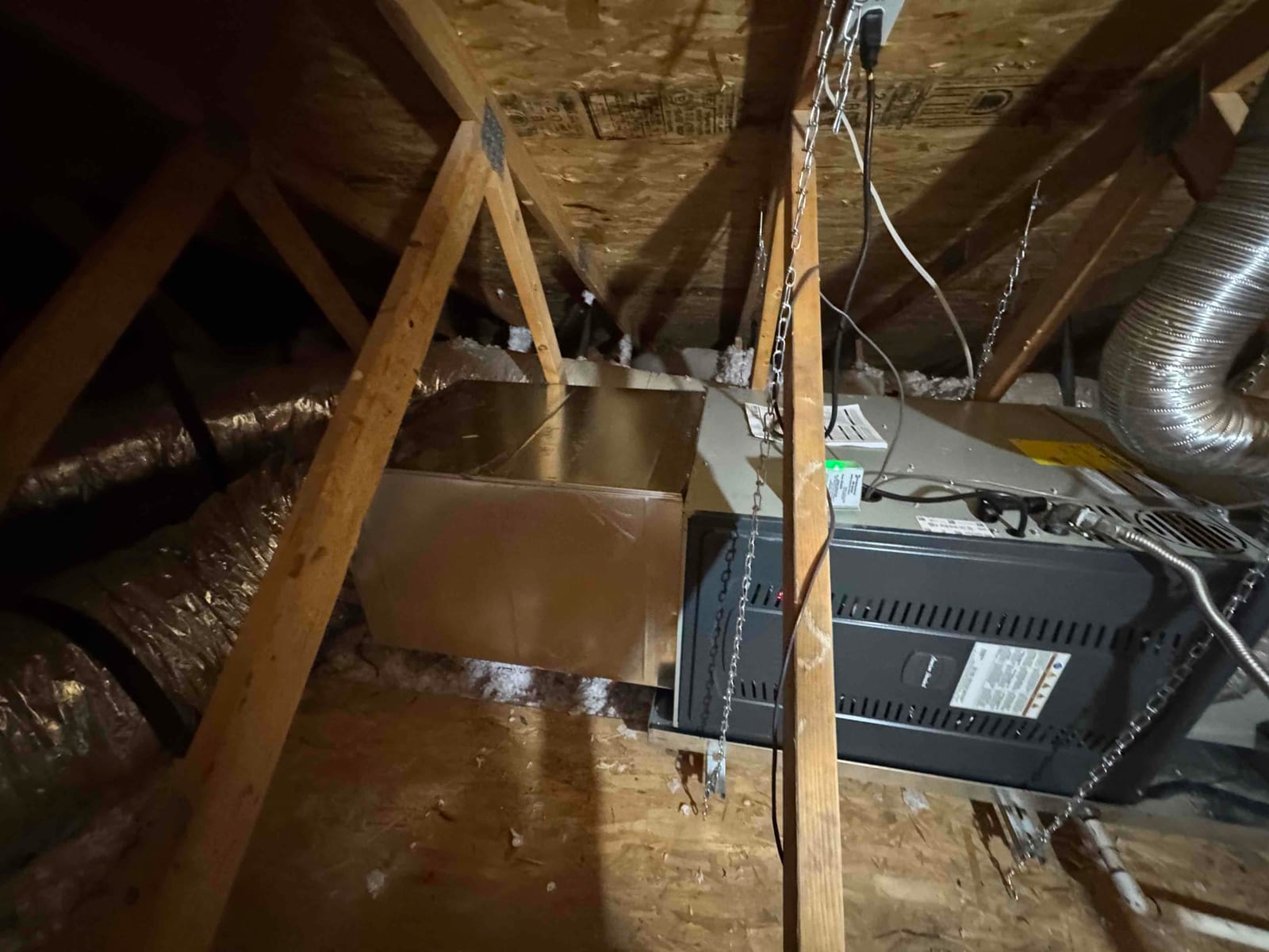 New attic air handler suspended with chains, connected to insulated ductwork and sealed metal plenum on a plywood platform.