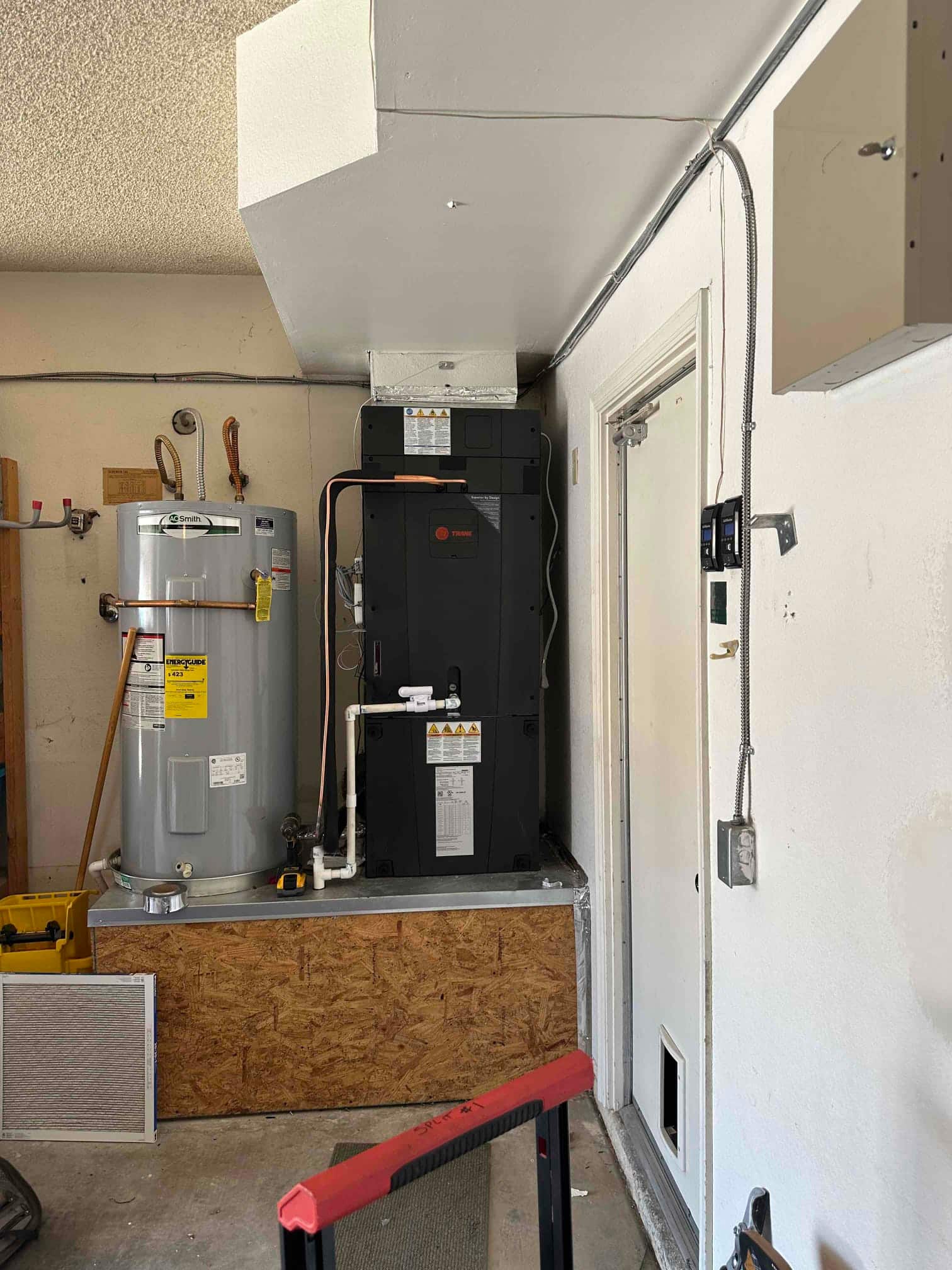 New indoor air handler installed on a raised platform beside a water heater, with condensate drain and insulated refrigerant lines connected.
