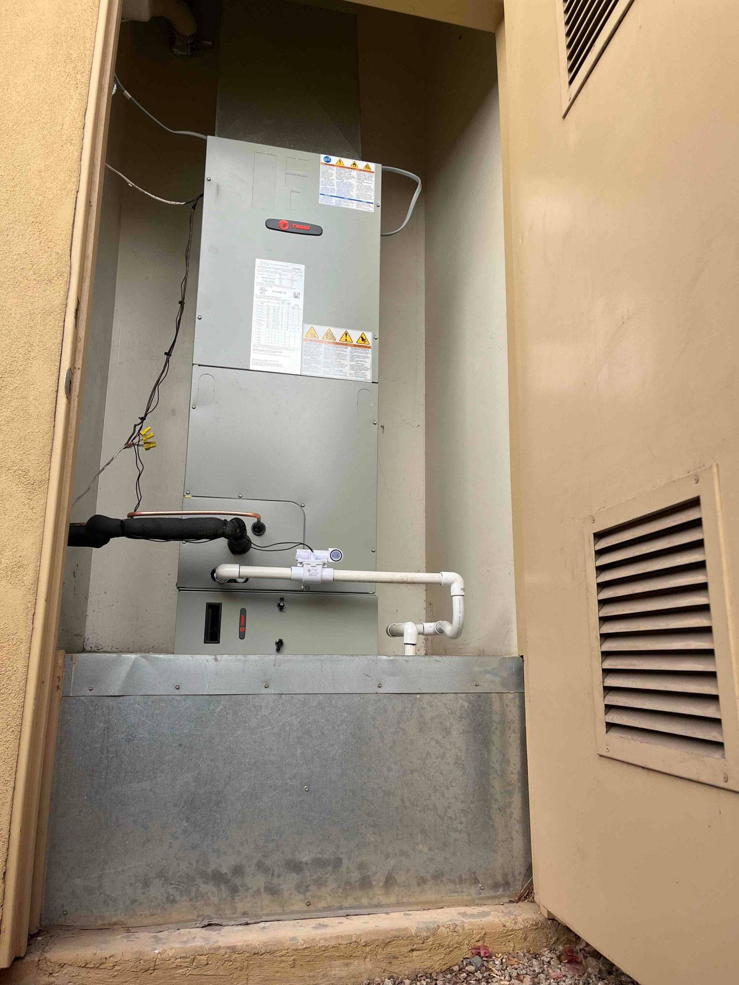New indoor air handler installed on a raised platform with insulated refrigerant line, PVC condensate drain, and connected ductwork in a recessed alcove.