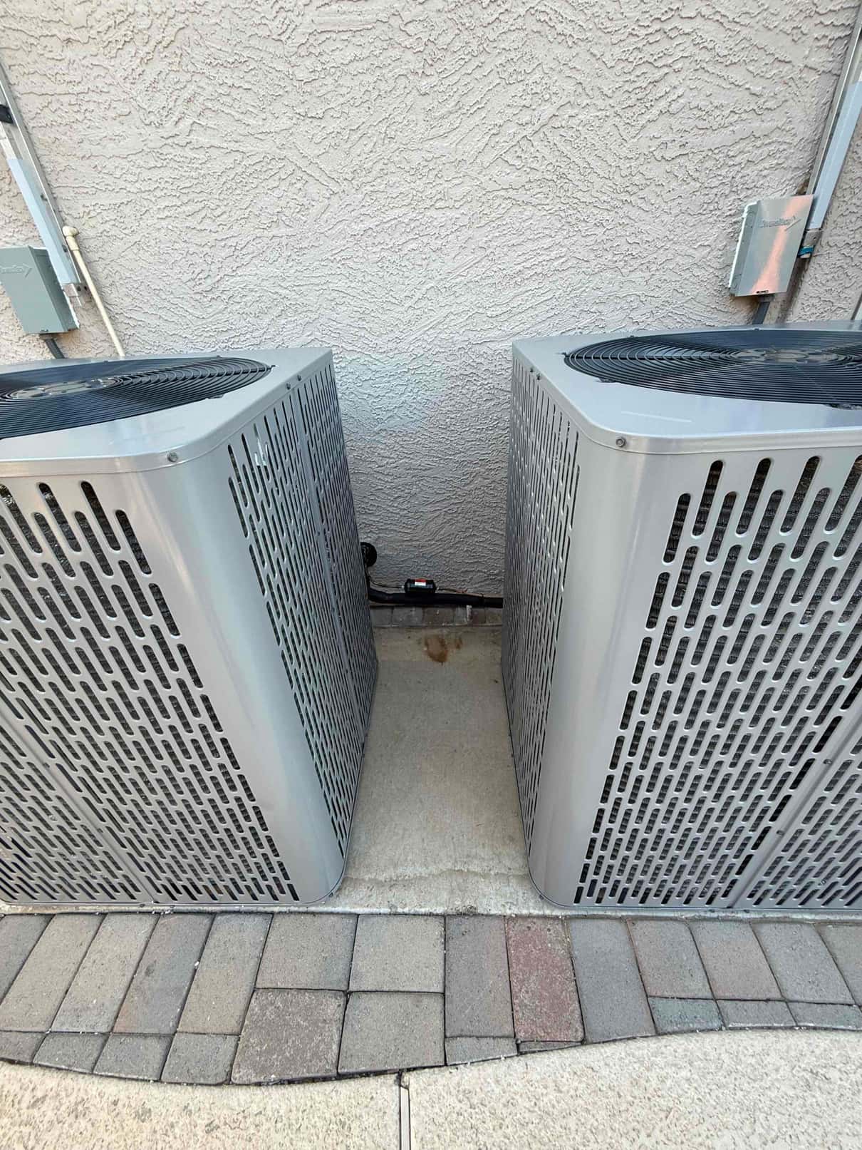 Two new outdoor condenser units set on a level pad with insulated refrigerant lines and electrical whip connections routed along the wall.