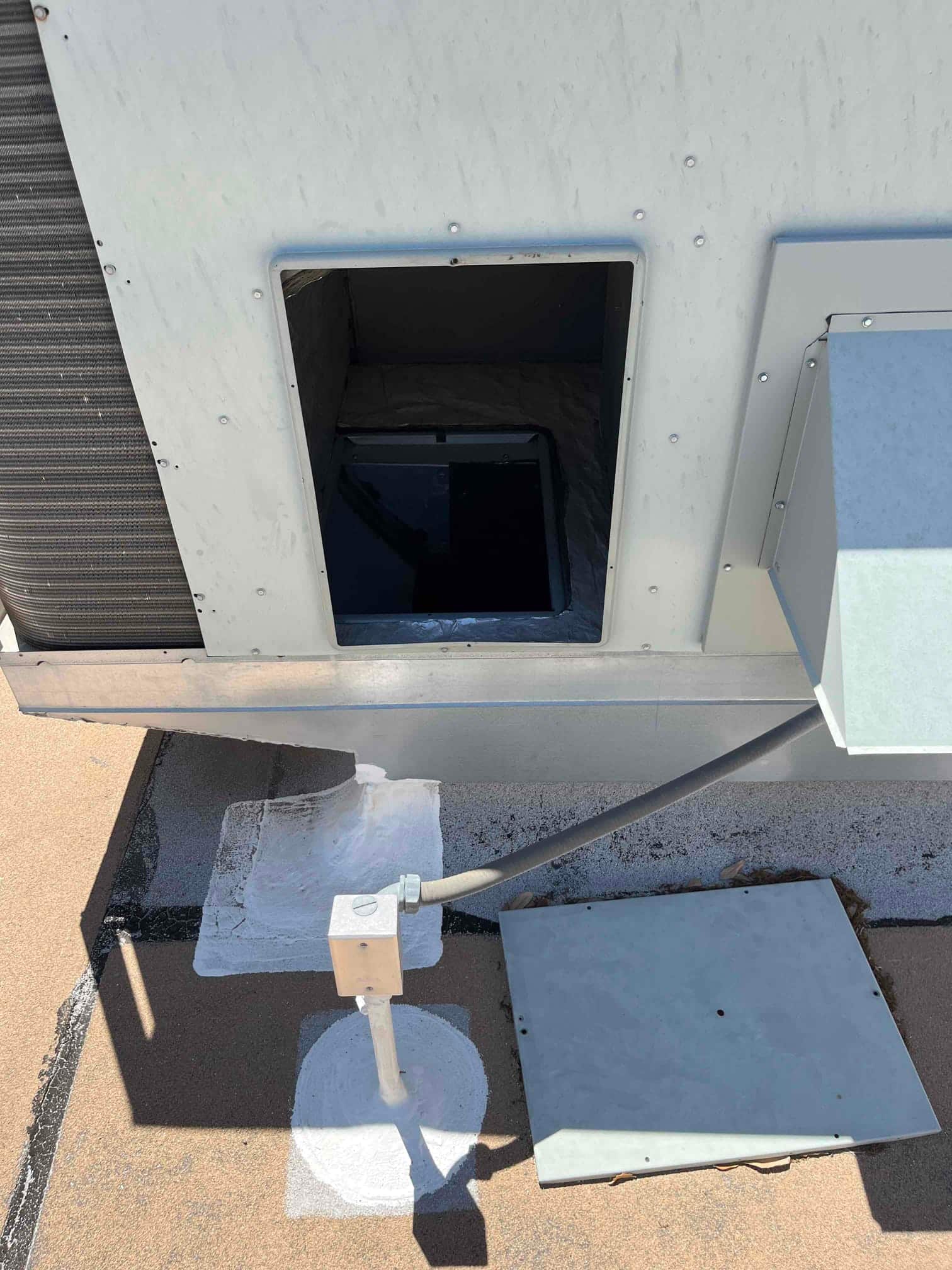 Open rooftop air handler access panel exposes interior compartment, with nearby electrical junction box and removed cover on the roof.