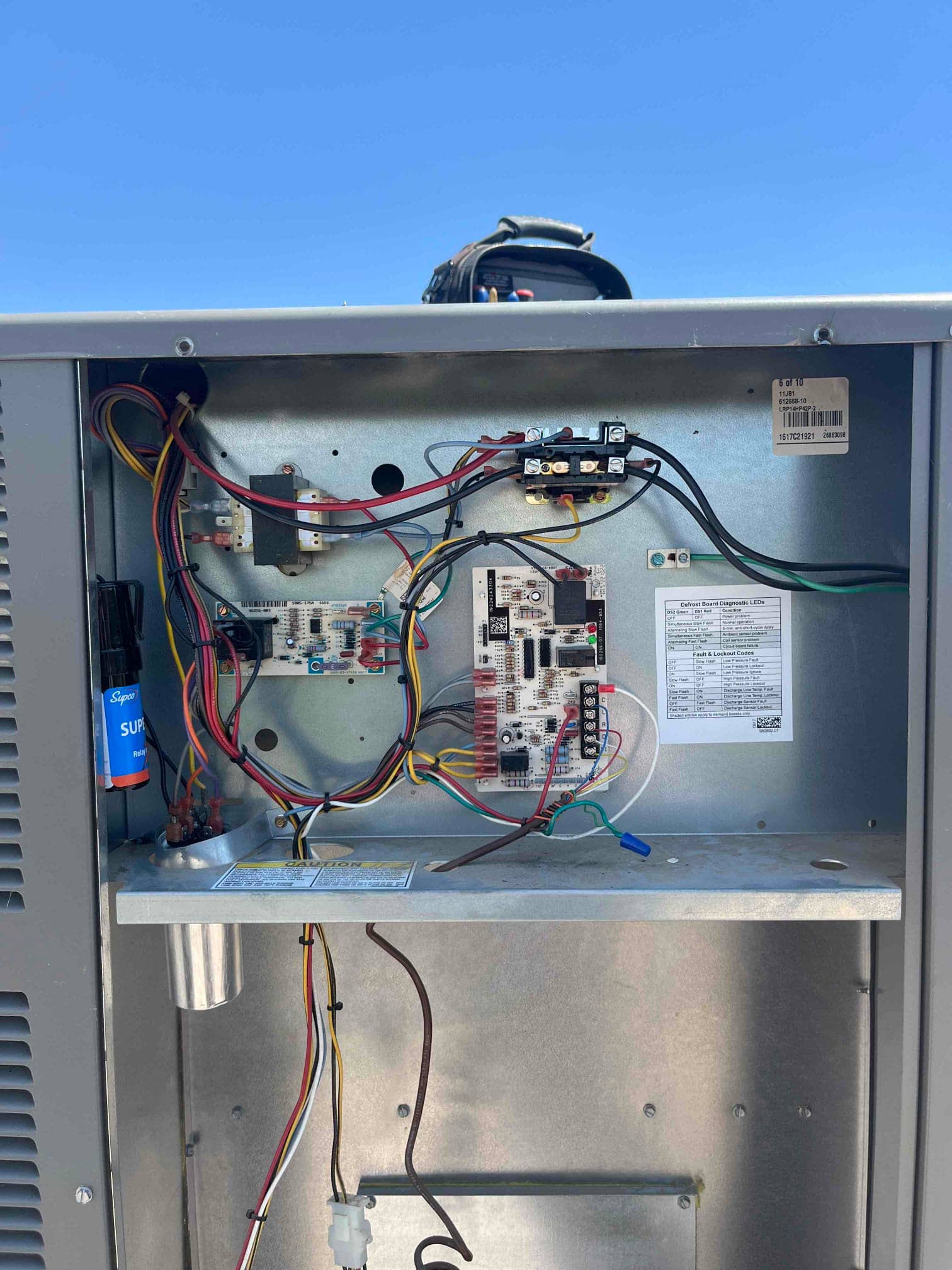 Outdoor condenser control compartment open, showing control board, wiring harnesses, inline fuse, and service label inside the panel.