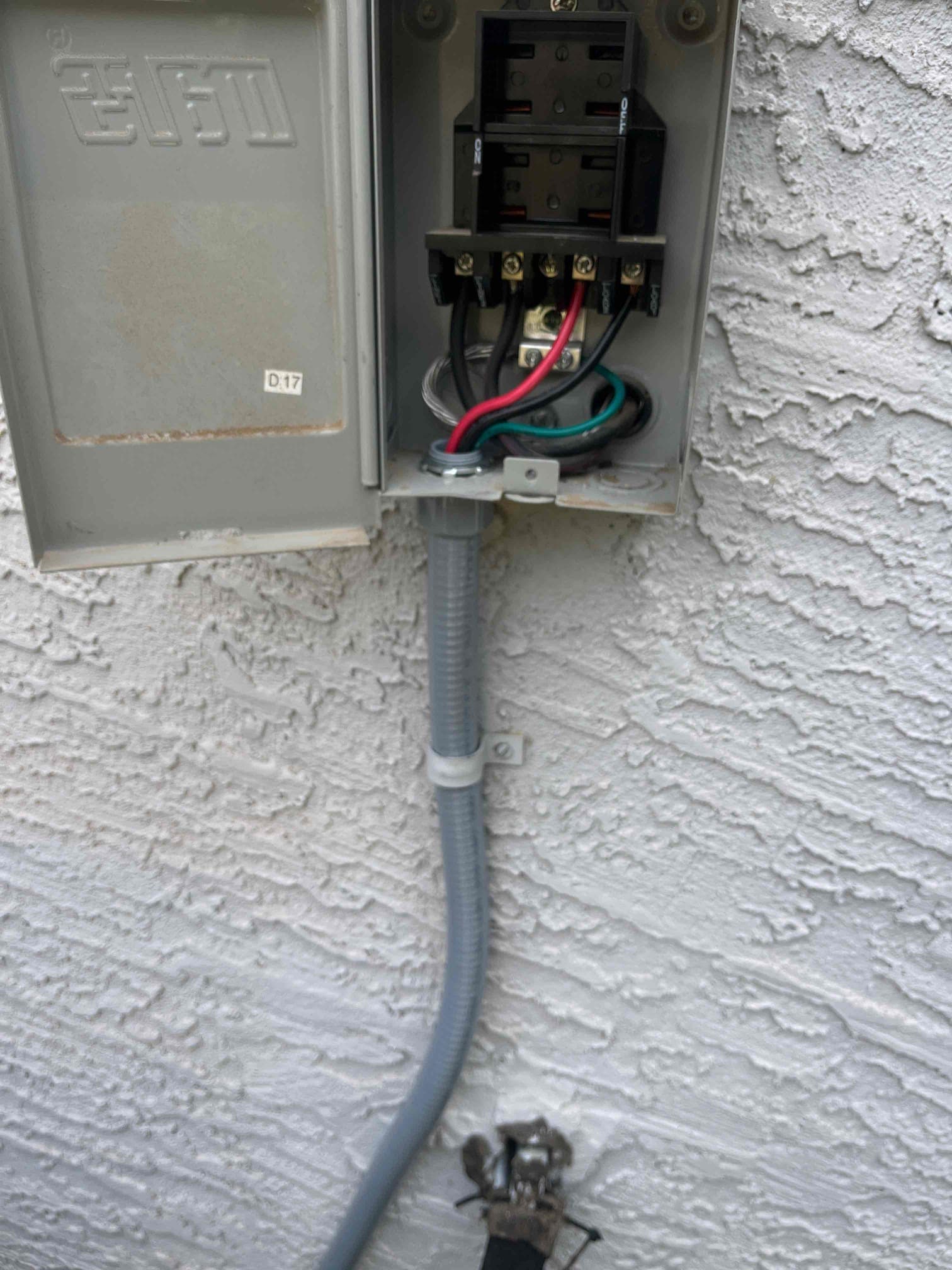 Outdoor disconnect box open, showing line and load wiring with flexible conduit and grounded bonding strap against stucco wall.