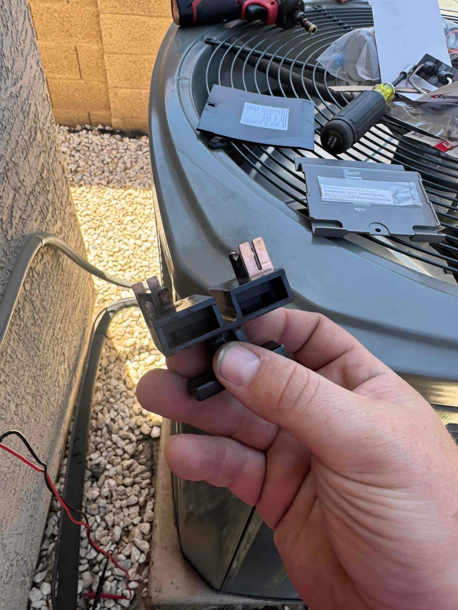 Technician holds a fused disconnect block with fuses removed beside an outdoor condenser unit during air conditioning repair.