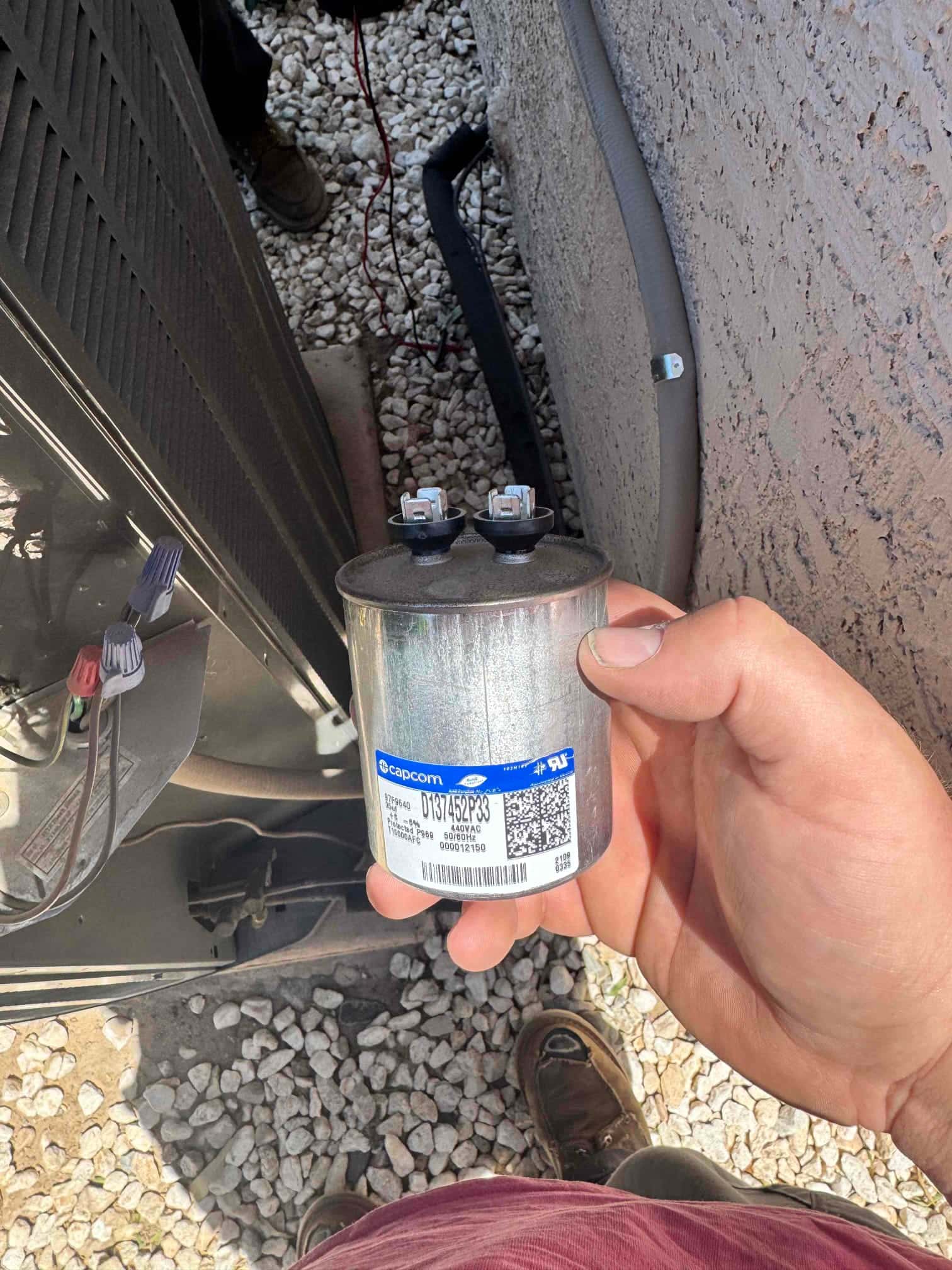 Technician holds a new dual-run capacitor beside the outdoor condenser, showing clean terminals and intact casing ready for installation.