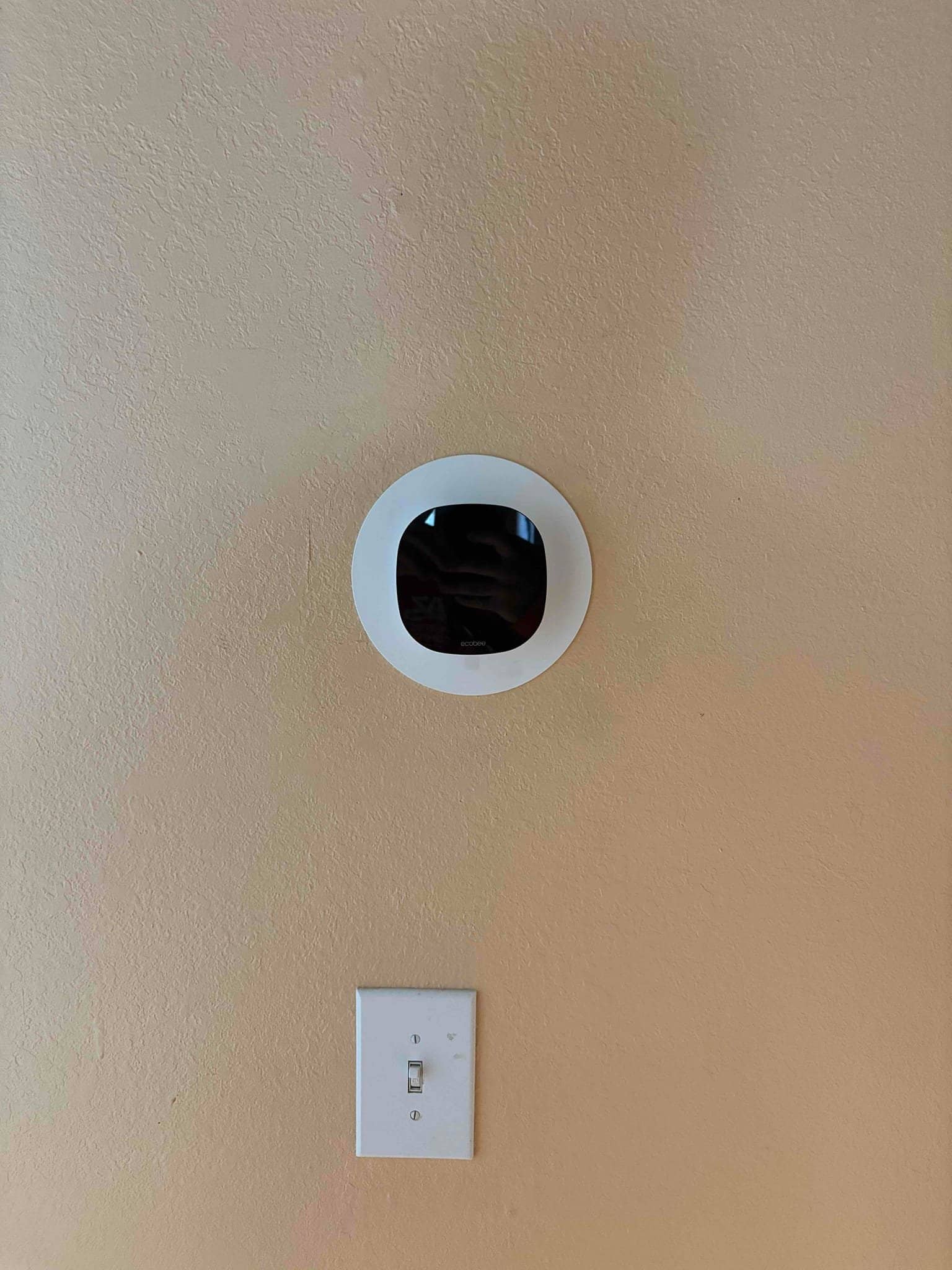 Wall-mounted smart thermostat installed with trim plate above a nearby light switch on a finished interior wall.