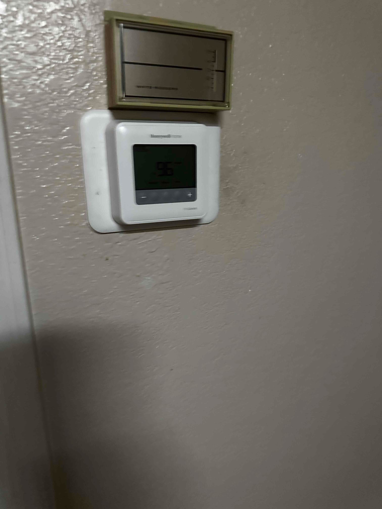 Wall-mounted thermostat installed on a backplate beneath an older control, showing a digital display and adjustment buttons.