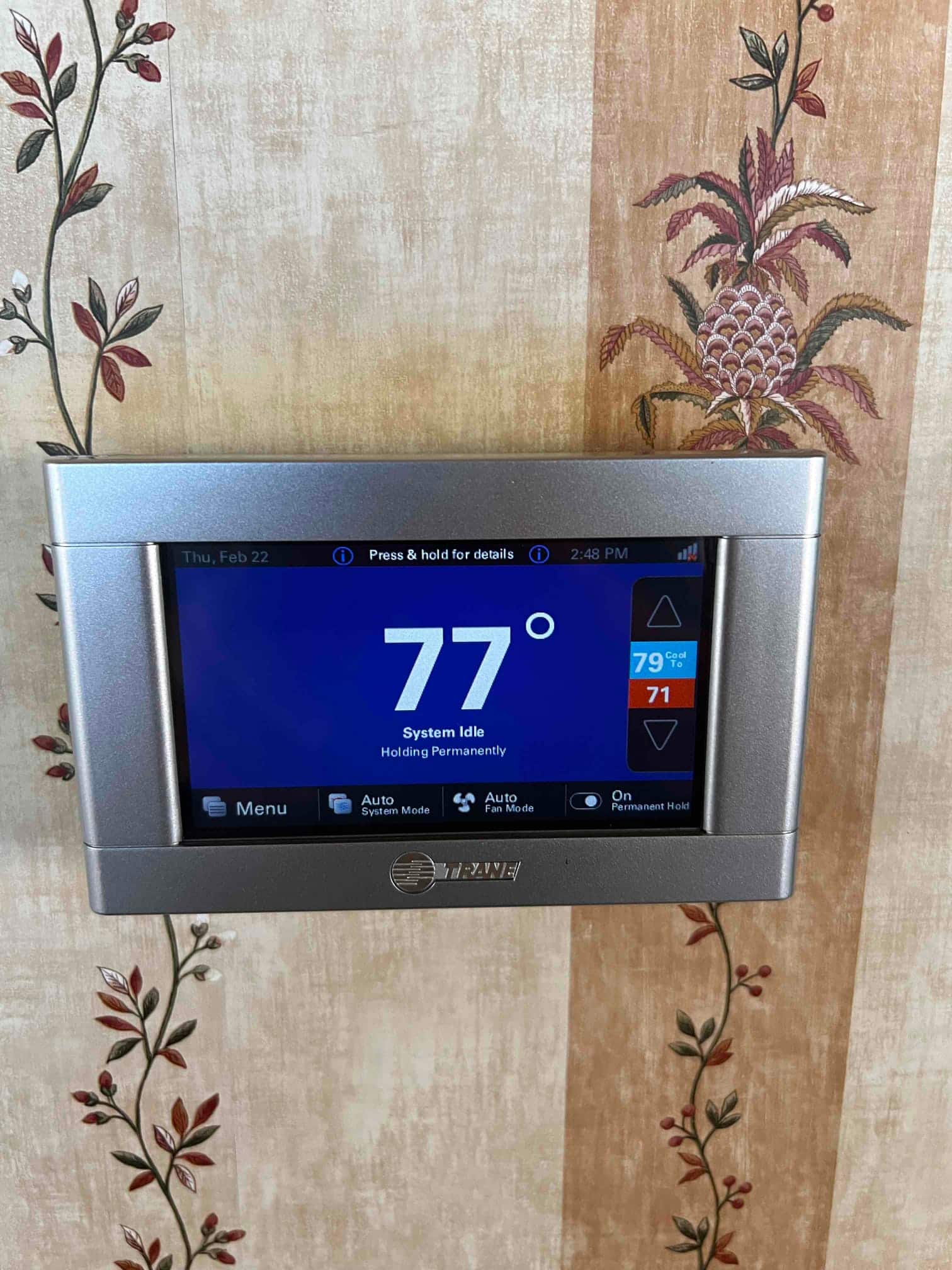 Wall-mounted touchscreen thermostat displays indoor temperature at 77 degrees with system idle and settings shown along the bottom.