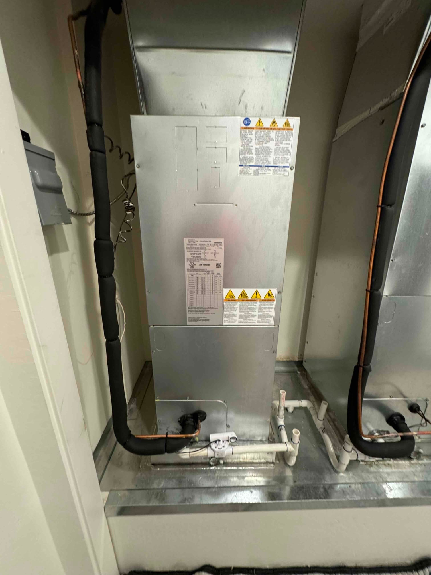 Indoor air handler installed on a drain pan with insulated refrigerant lines and PVC condensate piping connected and supported.
