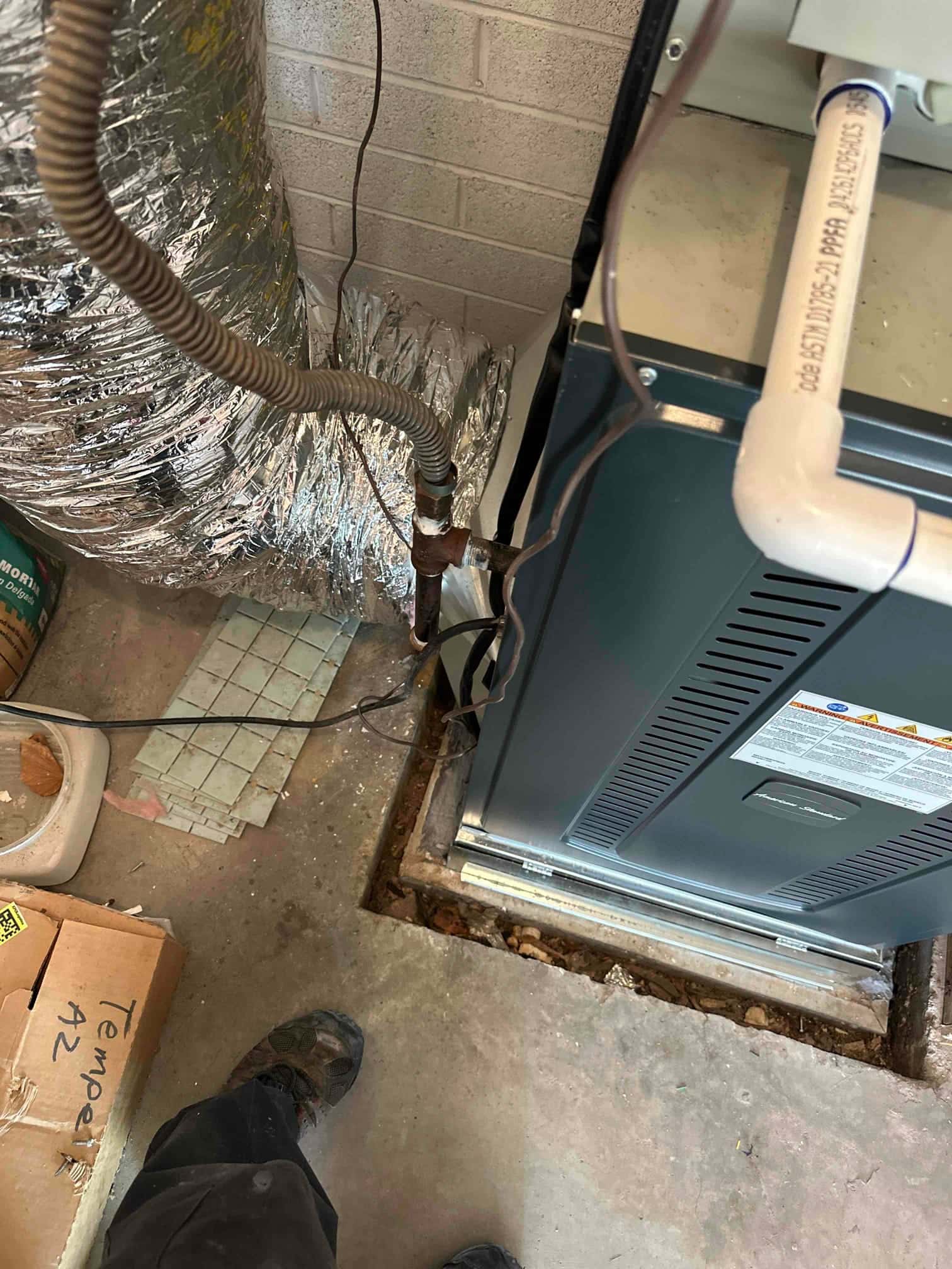 New furnace set in a recessed slab opening with connected PVC condensate drain and insulated flexible ductwork routed to the unit.
