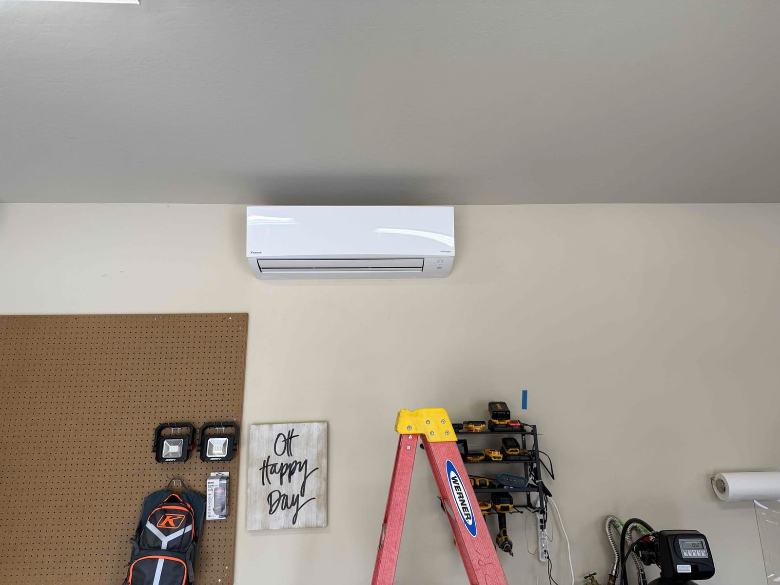 Wall-mounted mini-split indoor unit installed level on a finished wall above a workspace, with ladder and tools nearby.