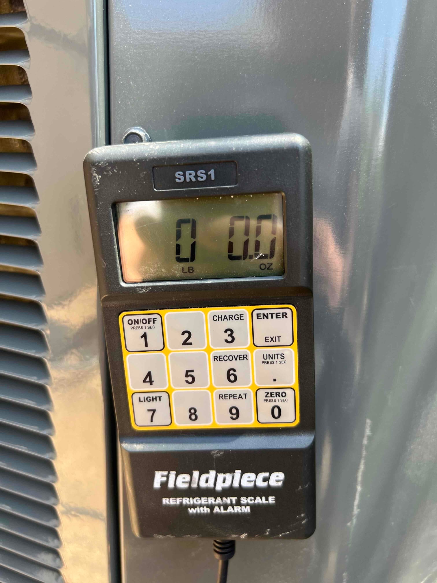 Digital refrigerant scale controller mounted on the outdoor condenser, display showing 0 lb 0.0 oz before charging operation.