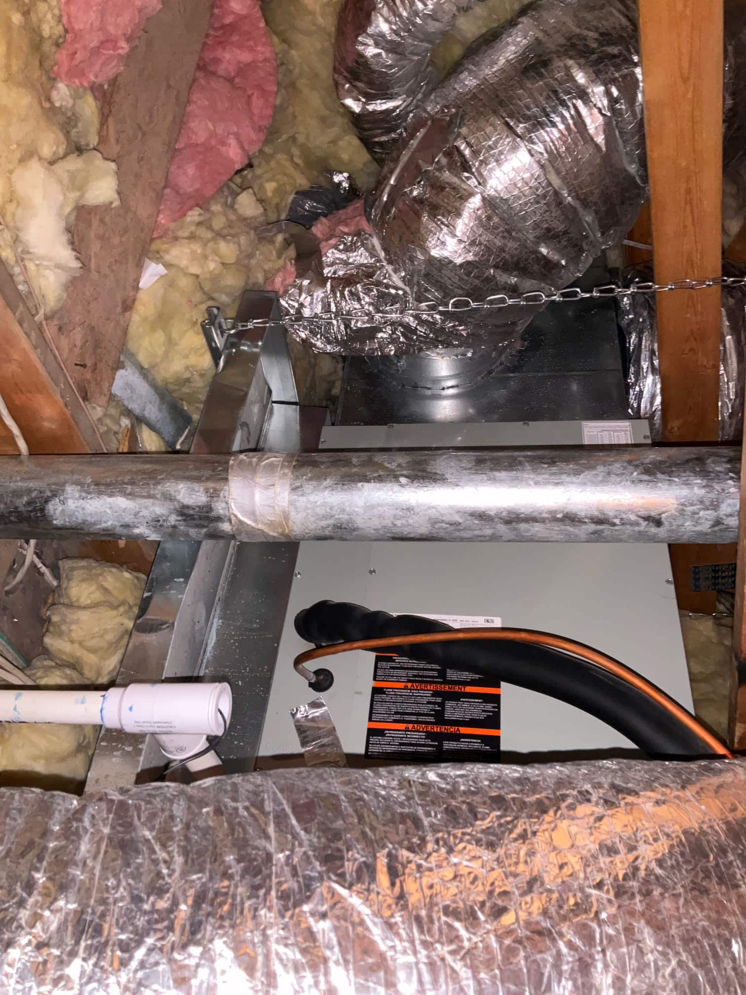 Attic air handler with connected flexible ducts, insulated refrigerant line set, and PVC condensate drain surrounded by fiberglass insulation.