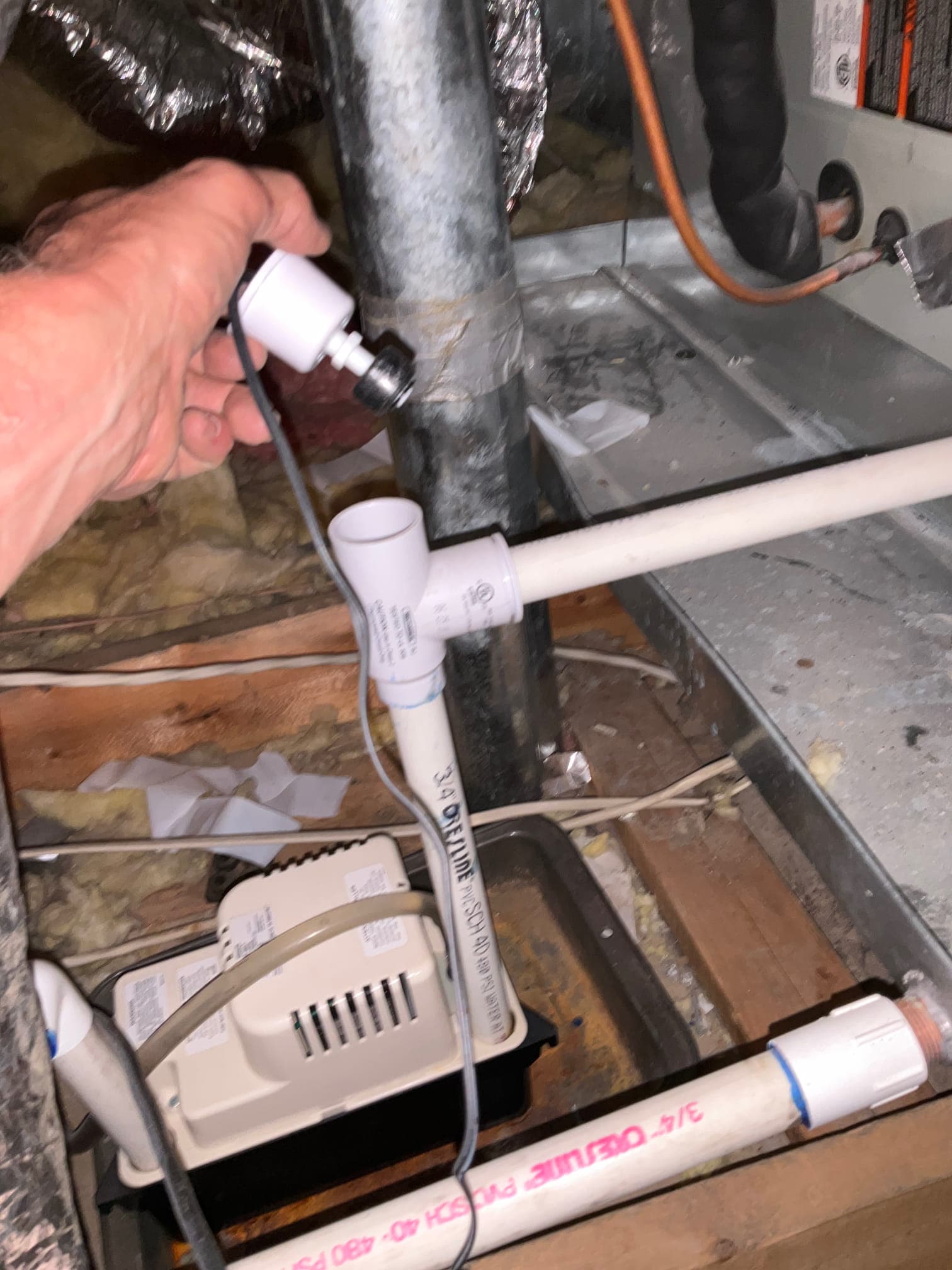 Condensate pump with PVC drain lines and overflow tee beside the air handler drain pan; hand holds a small wired sensor near ductwork.