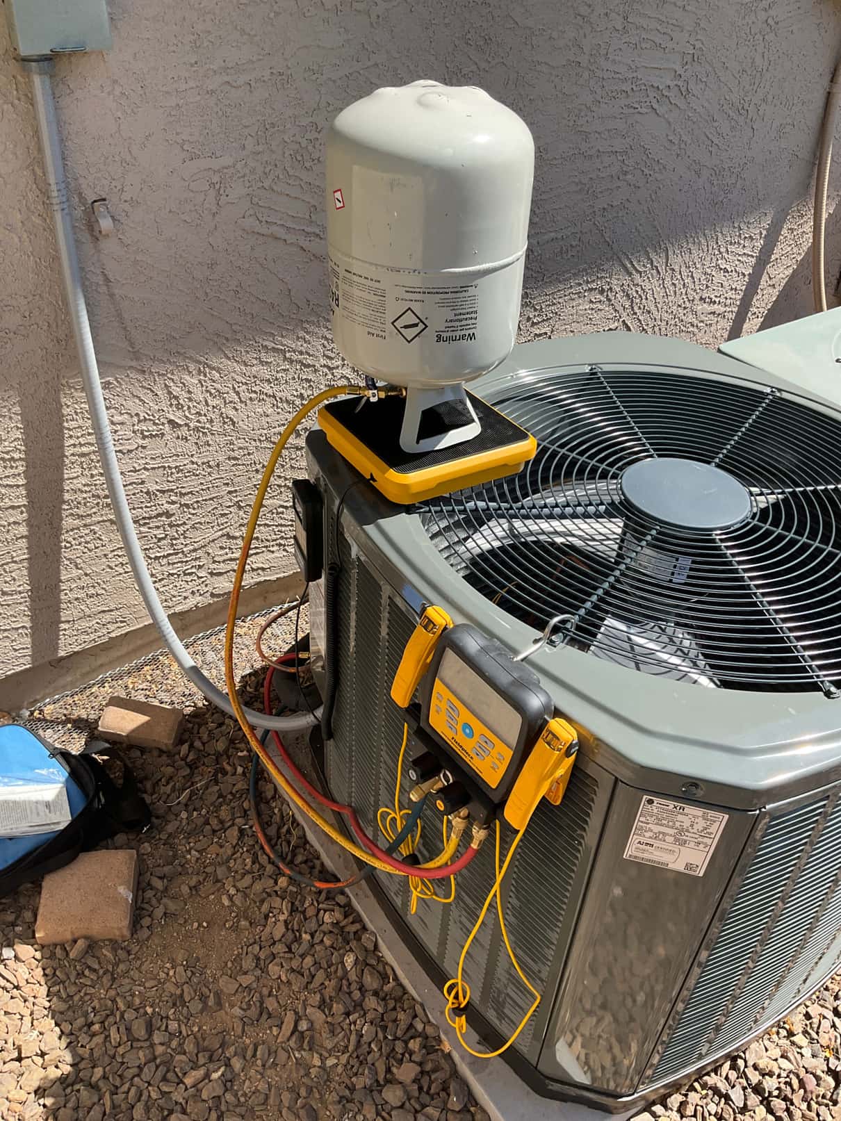 Digital manifold gauges connect to the new condenser while a refrigerant cylinder sits on a scale during charging.