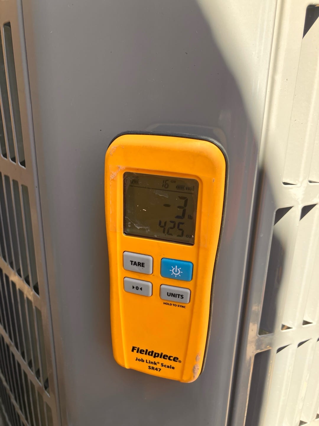 Digital refrigerant scale attached to the outdoor condenser during installation, showing a weight reading on its display.