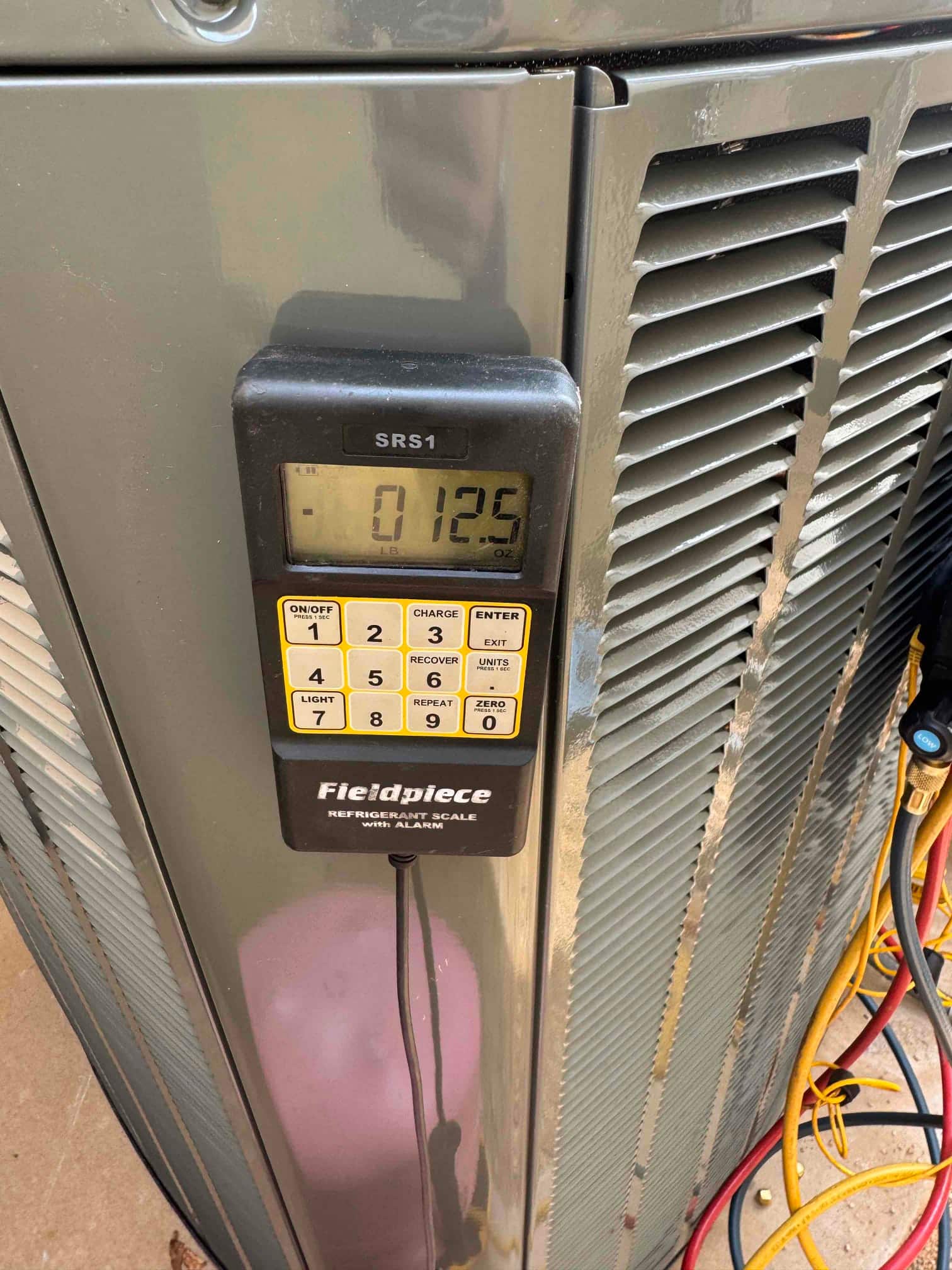 Digital refrigerant scale displays 12.5 ounces during charging beside the outdoor condensing unit with hoses connected.