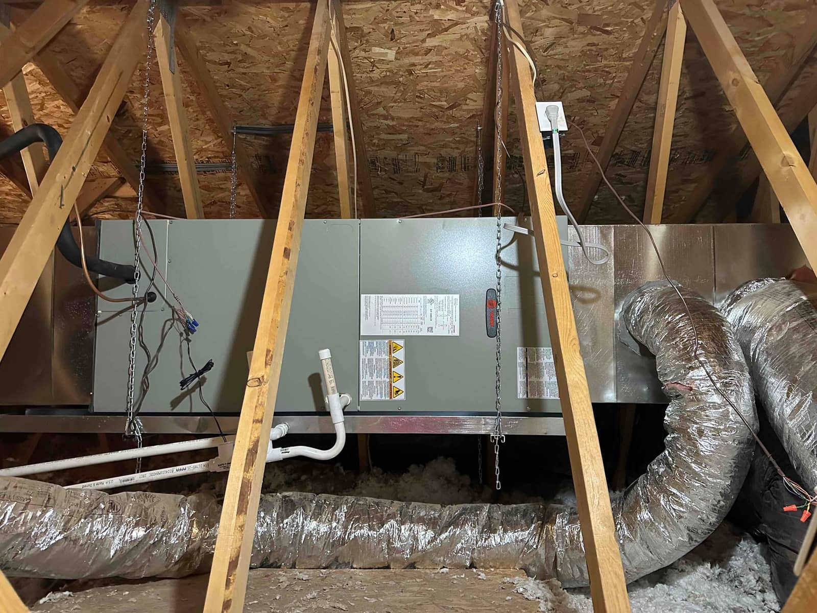 New air handler suspended in attic with insulated flex ducts connected, condensate drain piped, and electrical whip secured to a junction box.