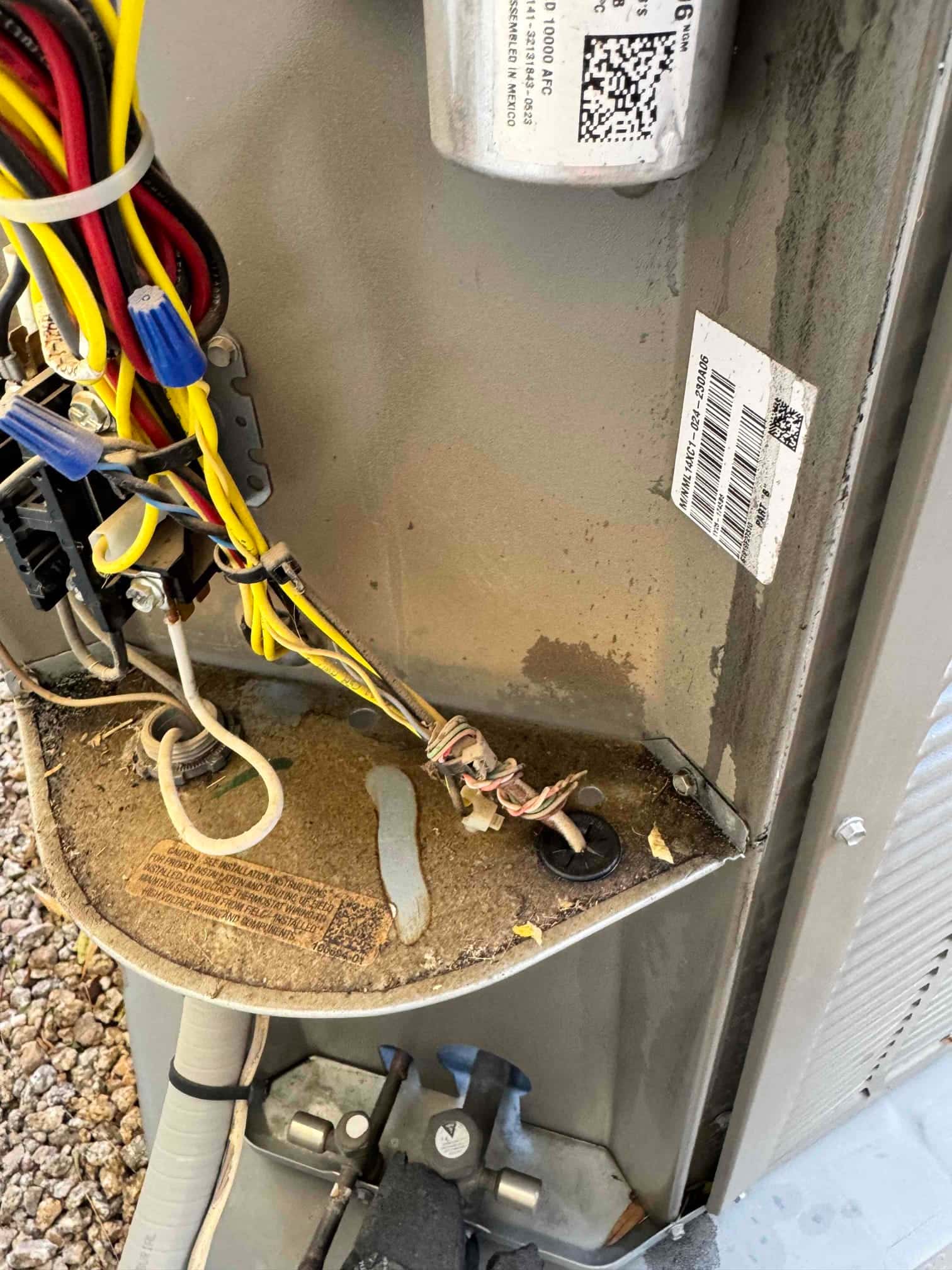 Outdoor condenser control compartment shows wiring, installed capacitor can, and dusty base panel with service labels and grommeted conduit.