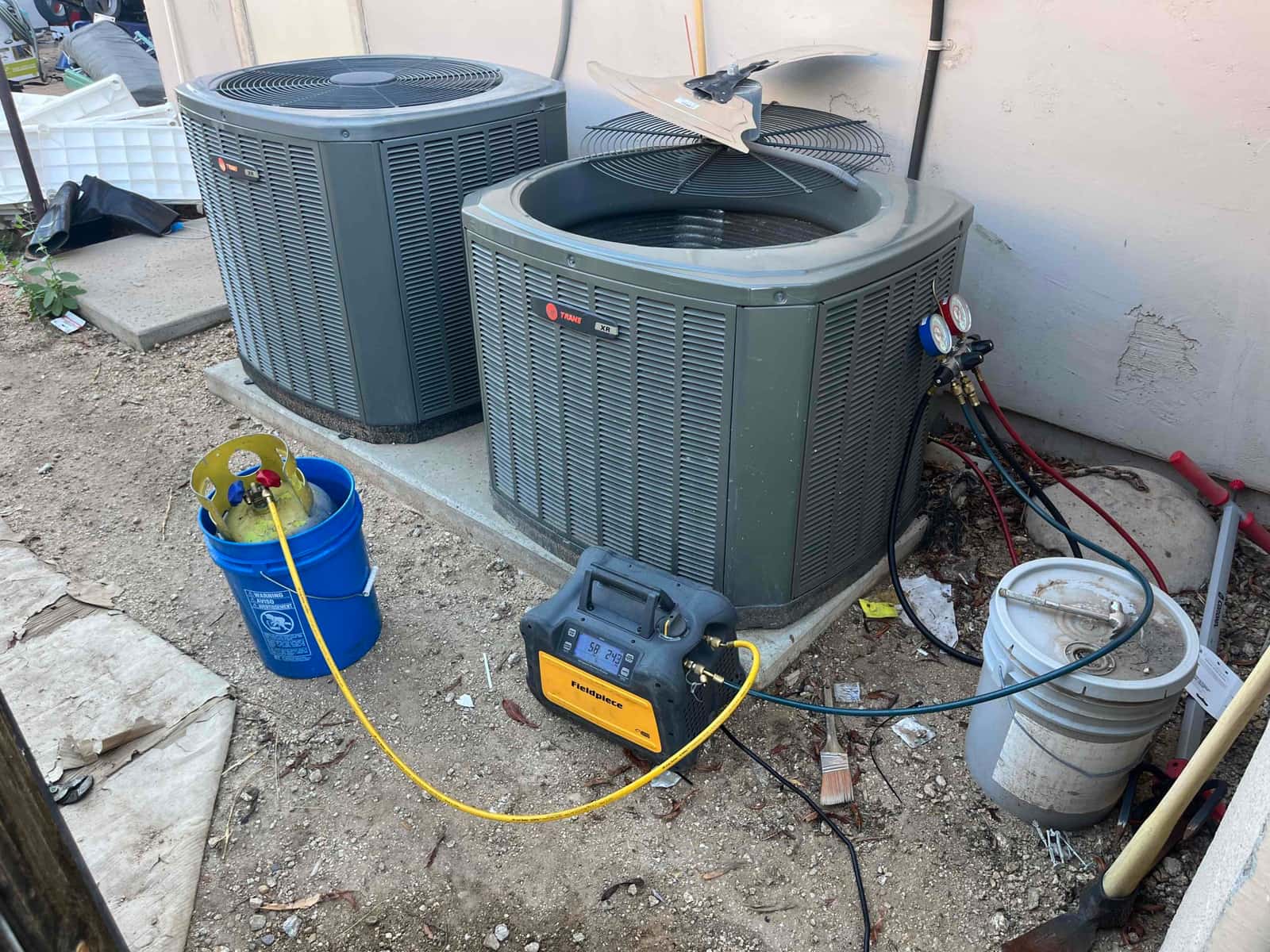 Two outdoor condenser units undergoing service with refrigerant recovery machine, recovery cylinder, and manifold gauges connected to the right unit.