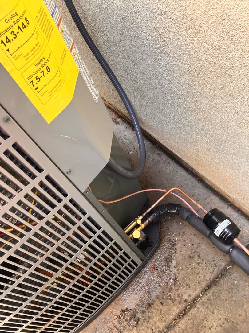 Outdoor heat pump condenser set beside wall with insulated refrigerant lines, service valves, and a liquid-line filter-drier installed.