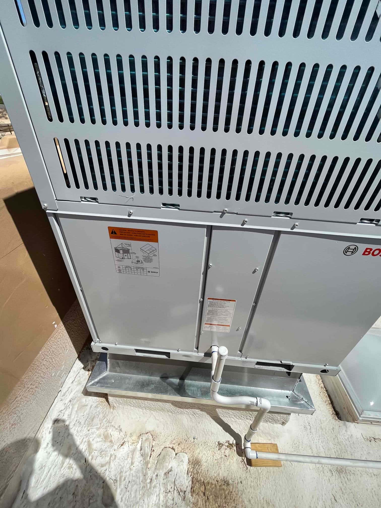 Outdoor heat pump placed on a metal stand with routed PVC condensate drain and sealed roof surface around the unit.