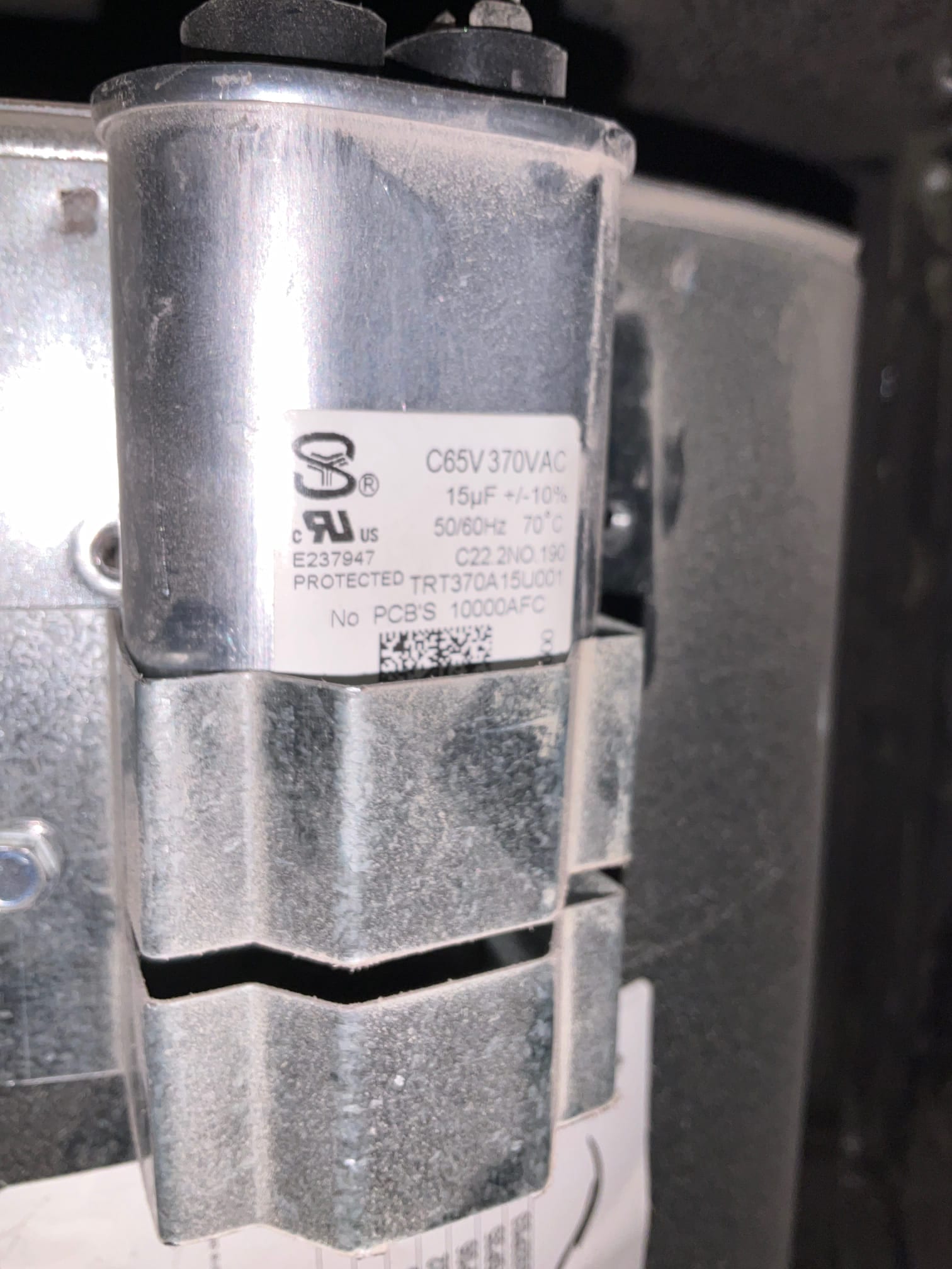 Oval run capacitor labeled 15 μF 370VAC, mounted with a metal strap to a galvanized housing.