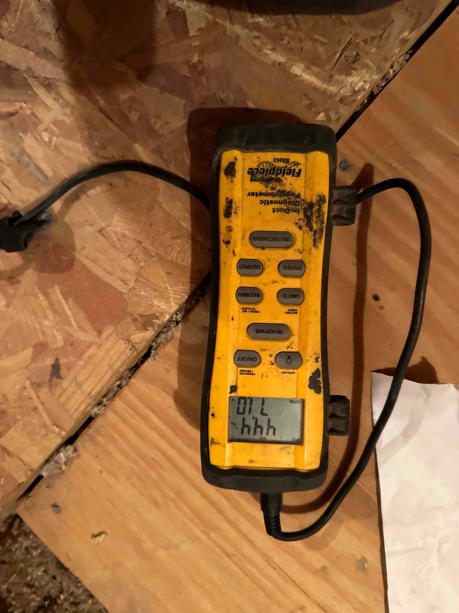 Portable diagnostic meter rests on plywood subfloor with display active and control buttons visible, powered by attached cable.