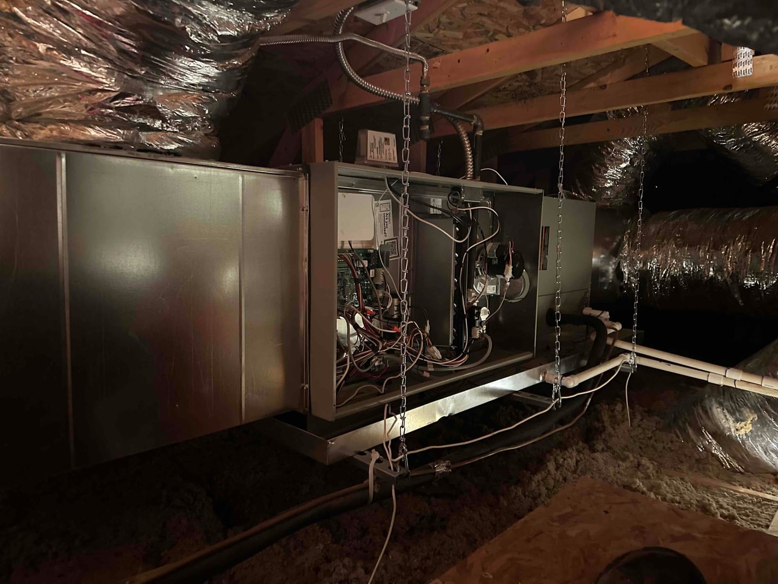 Suspended attic air handler installation with connected flexible ducts, condensate drain piping, and service panel open for wiring access.