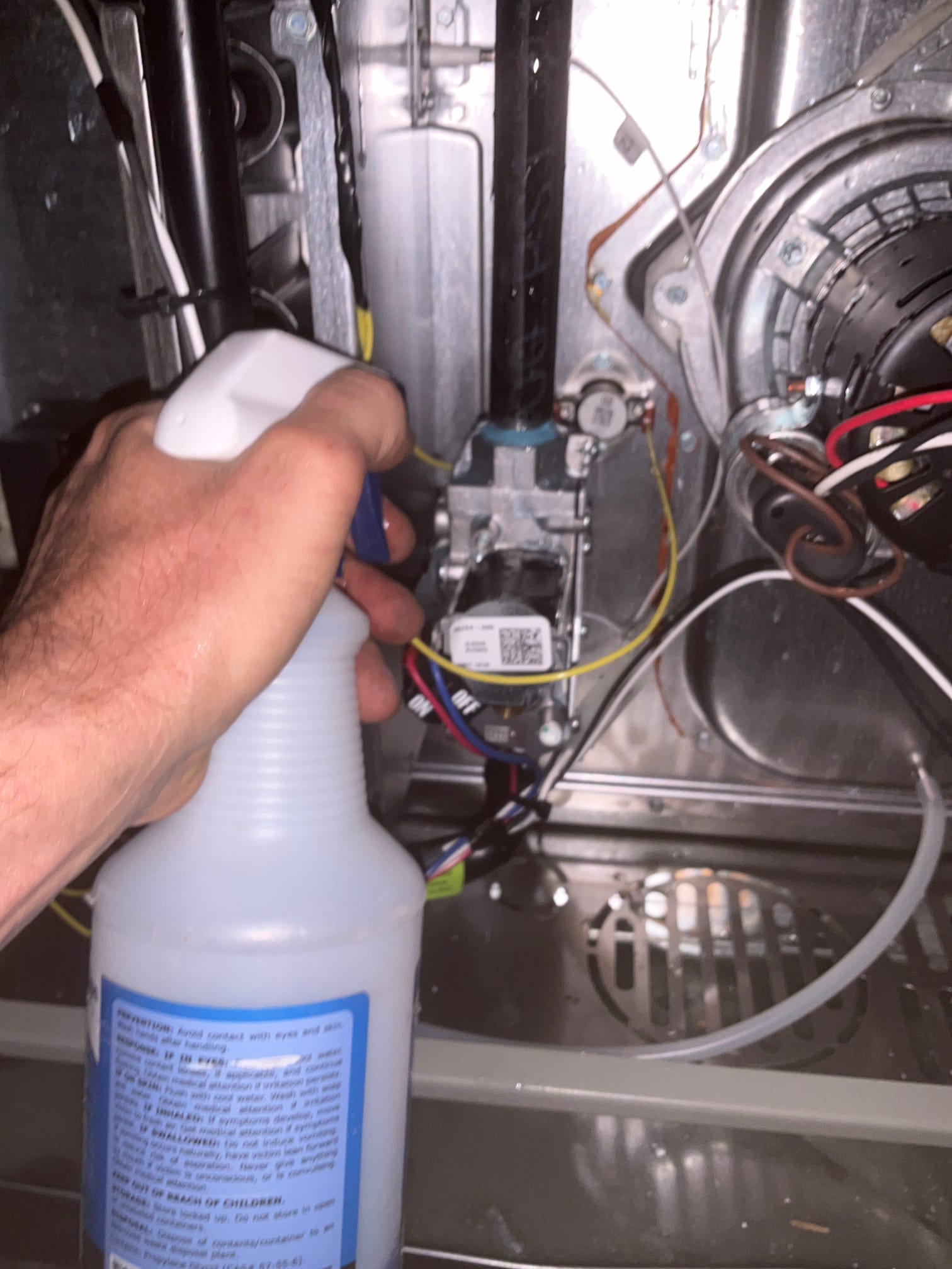 Technician sprays cleaner inside the furnace cabinet near wiring, gas valve, and inducer assembly during installation cleanup.