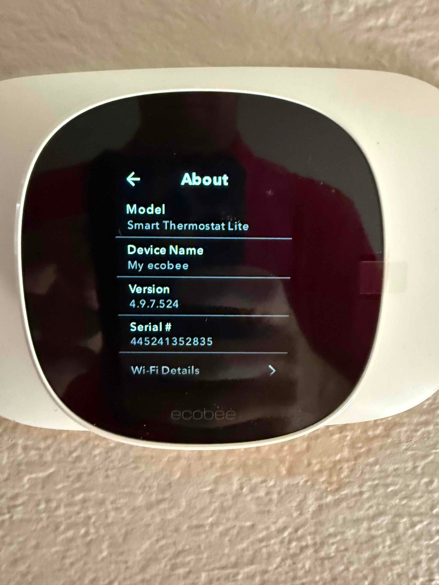 Wall-mounted smart thermostat displays the About screen with model, device name, software version, and Wi‑Fi details menu.
