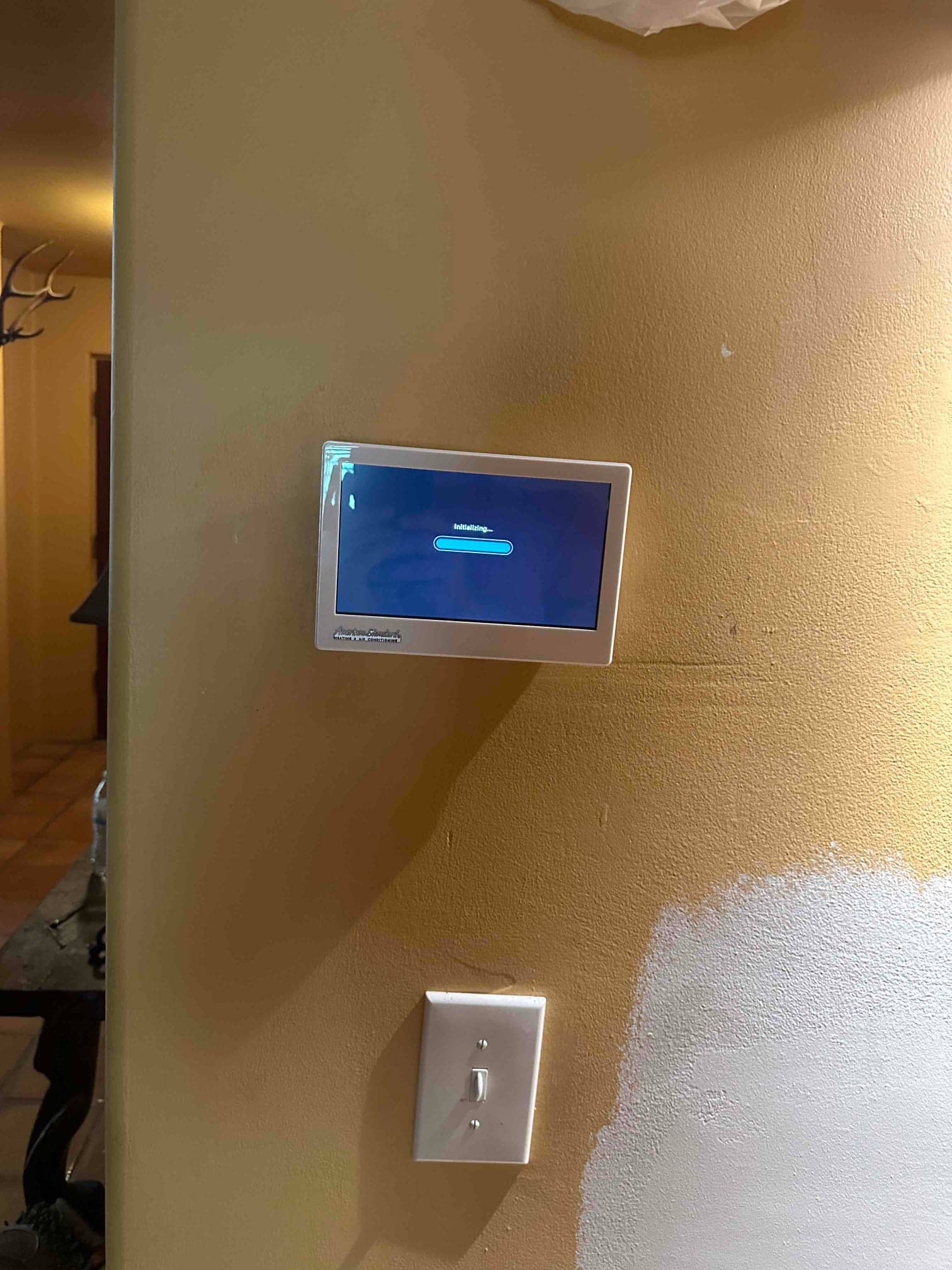 Wall-mounted smart thermostat powers on with an initializing screen after installation, shown above a standard light switch.
