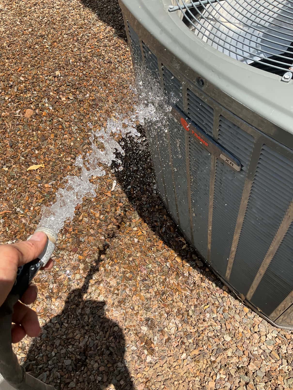 Water from a garden hose sprays across the side grille of an outdoor condenser.