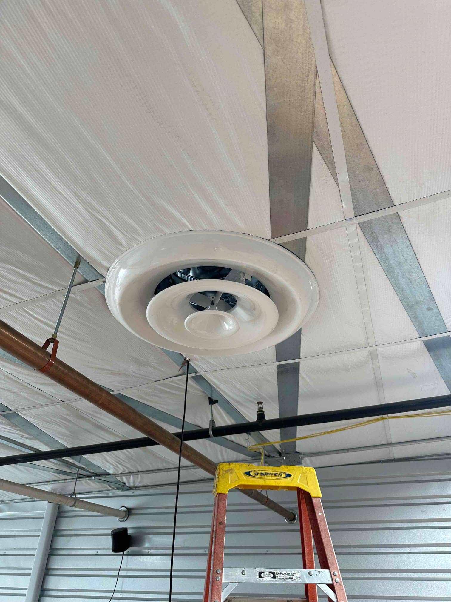 Ceiling-mounted round supply diffuser installed in insulated metal building ceiling, with nearby piping, hanger supports, and a ladder beneath.