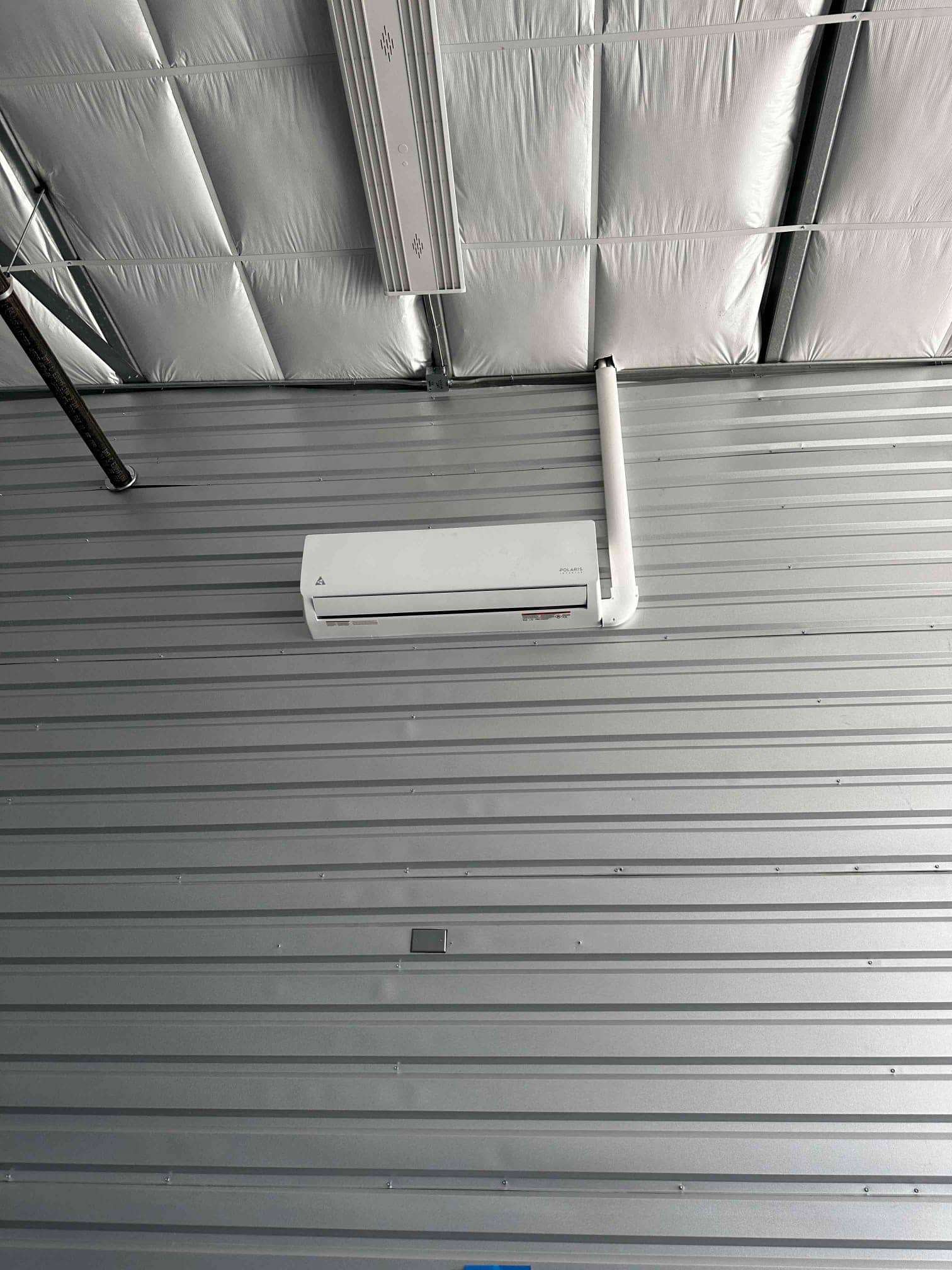 Wall-mounted mini-split indoor unit installed on a metal-clad wall with insulated ceiling above and condensate drain routed upward.