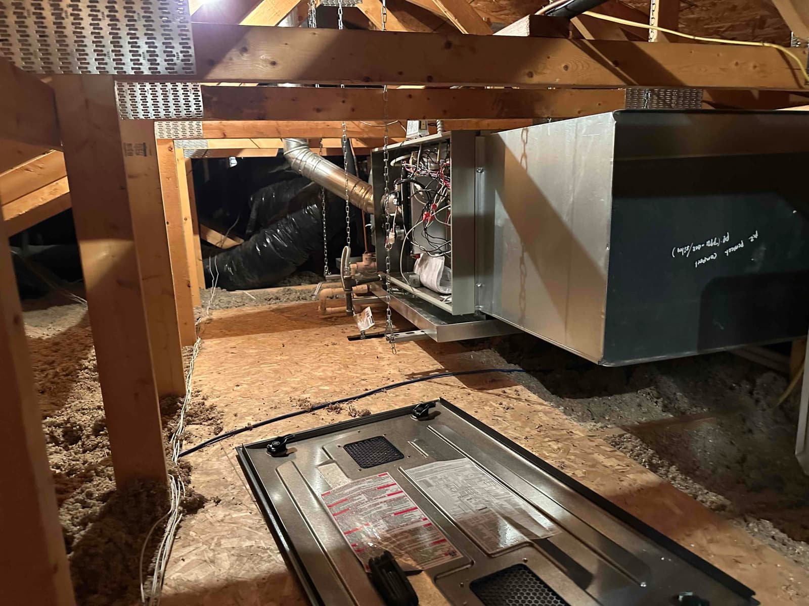 Attic installation showing a suspended air handler with connected ductwork, condensate piping, and service panel removed on the floor.