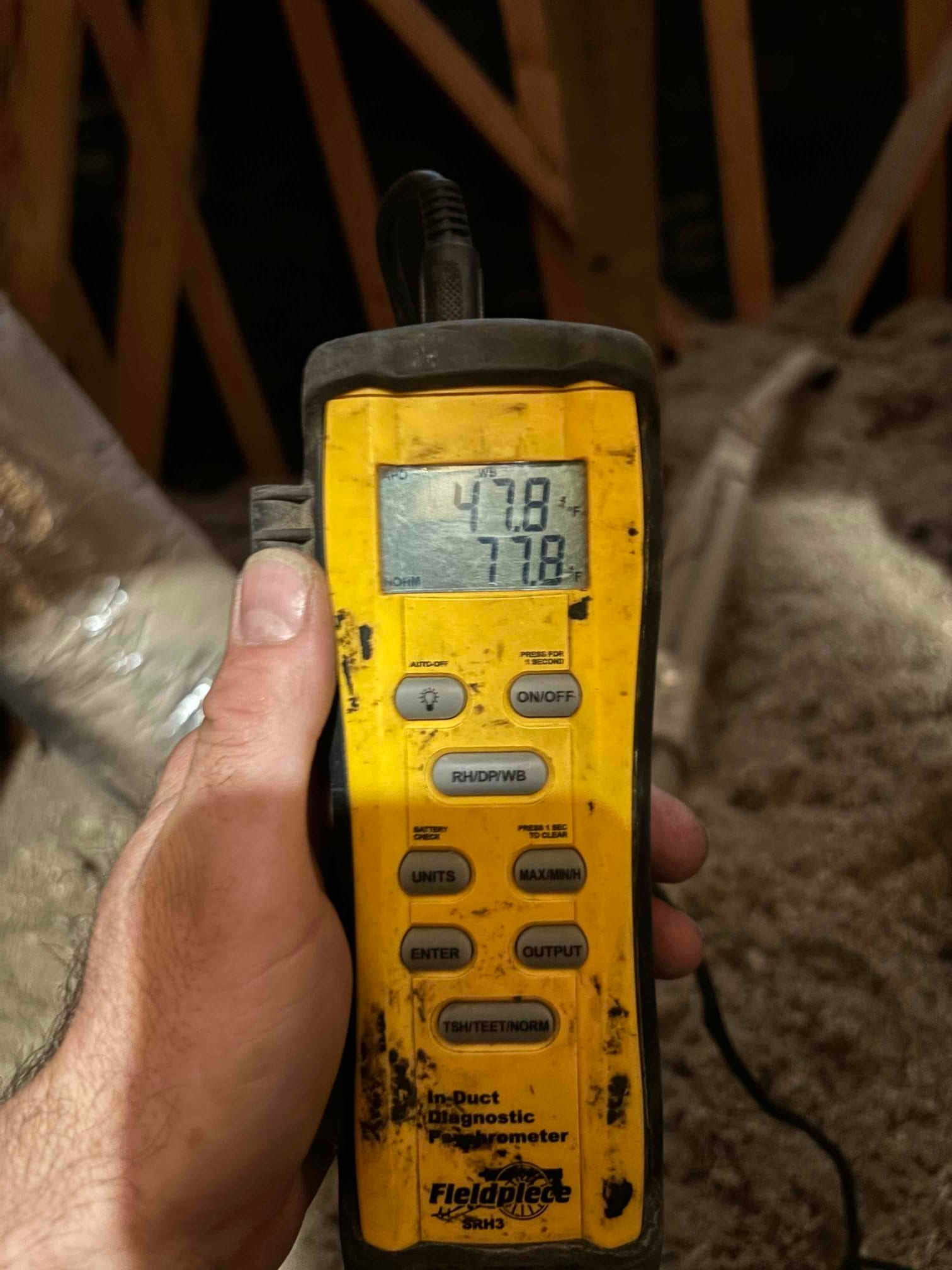 Handheld digital thermometer displays 47.8°F and 77.8°F during in-duct air temperature checks inside the attic.