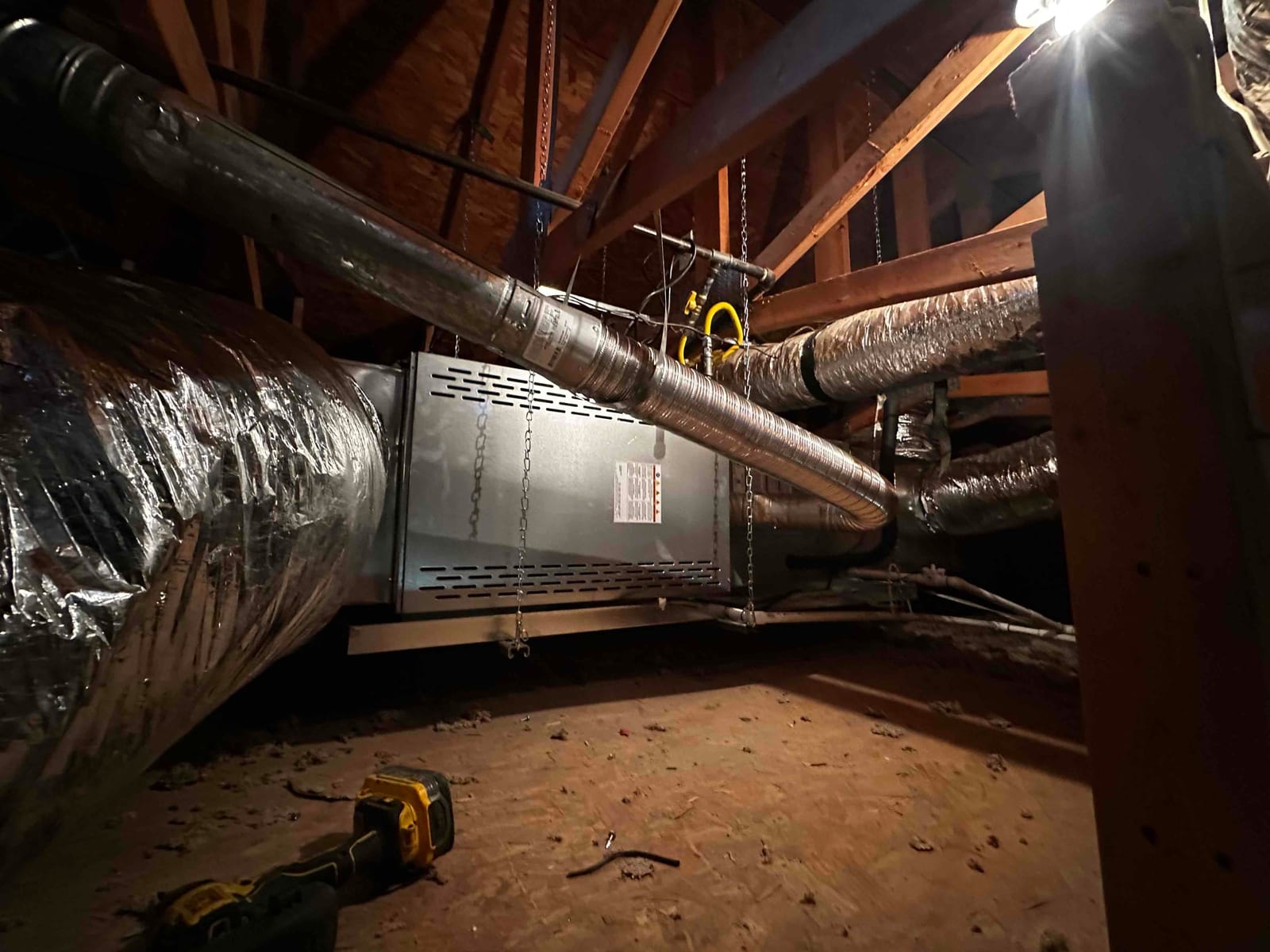 Horizontal air handler suspended in attic with connected flexible ducts, metal branch ducts, and support chains visible during installation.
