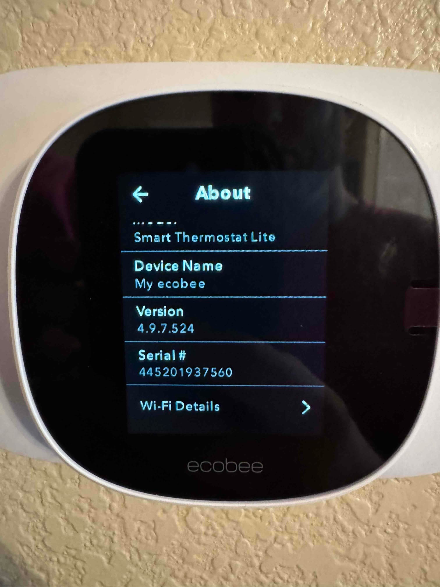 Smart thermostat mounted on wall displaying device information screen with software version and Wi‑Fi details menu.