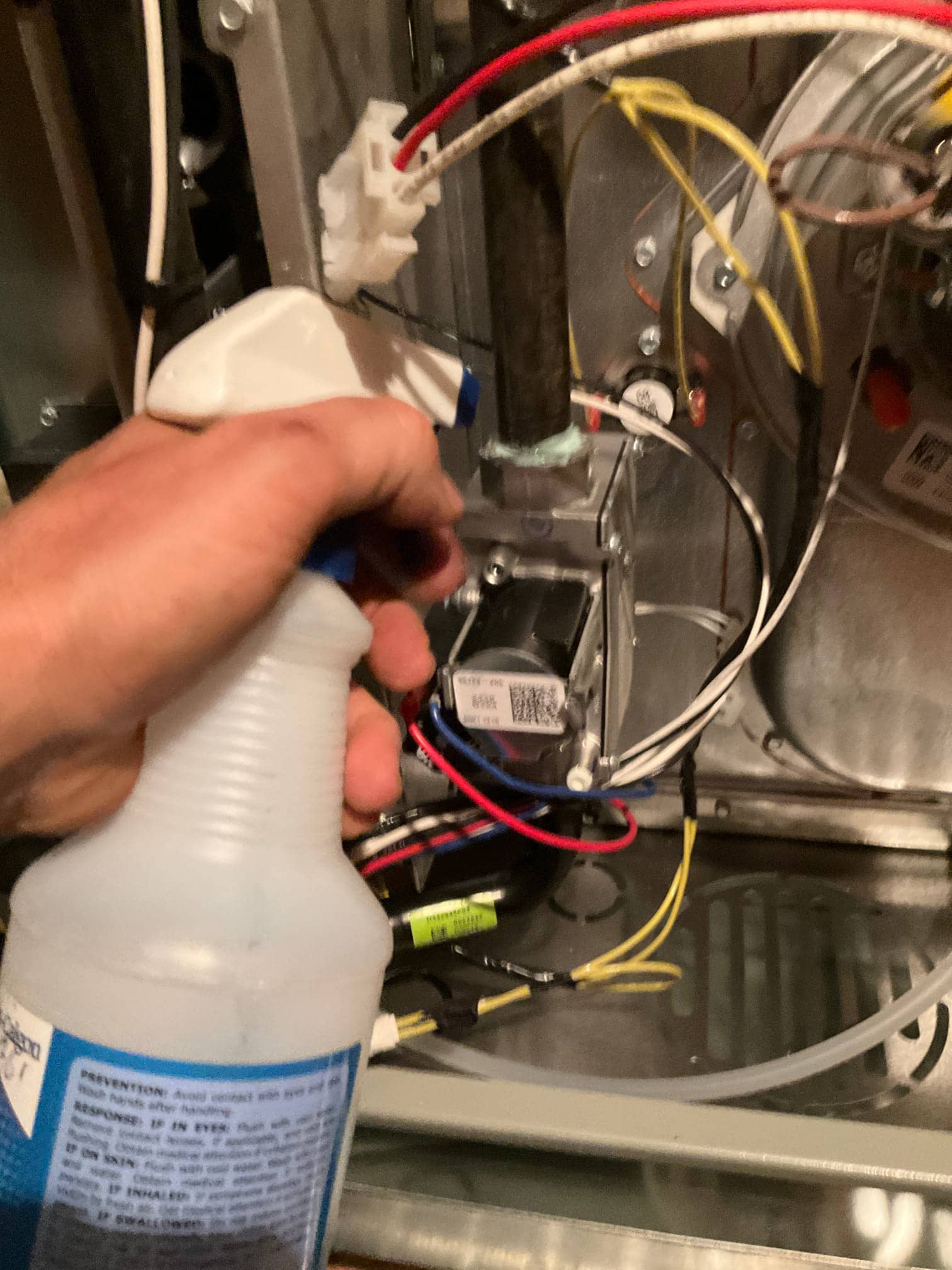 Technician sprays leak-detection solution on furnace cabinet penetrations and fittings during indoor unit installation, with wiring and components exposed.