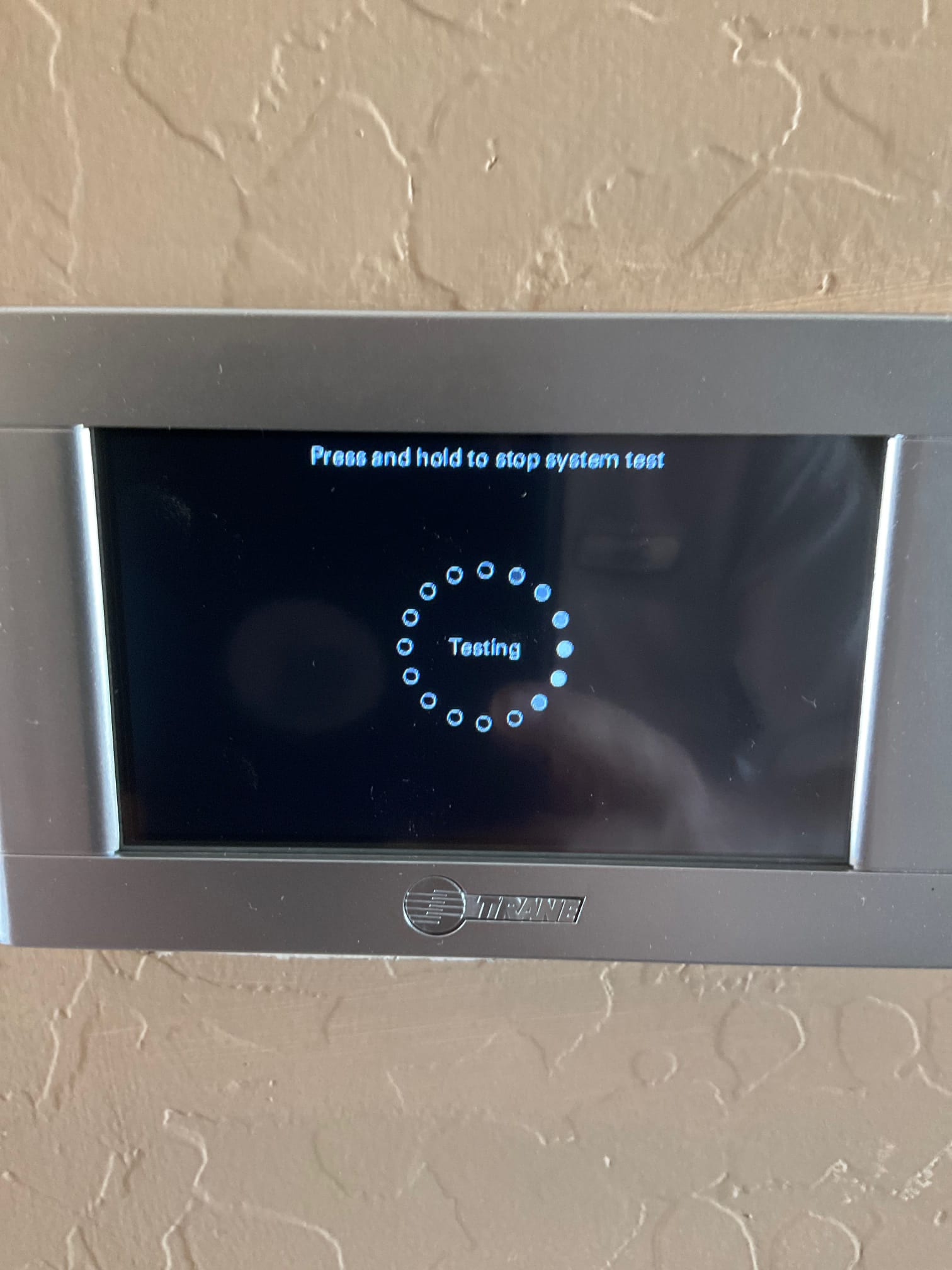 Wall-mounted touchscreen thermostat displays a system testing screen during commissioning after installation.