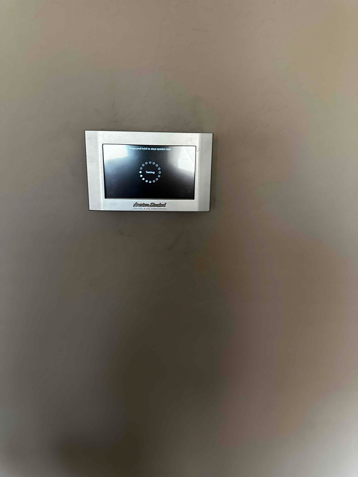 Wall-mounted touchscreen thermostat displays a system testing screen during startup after installation.