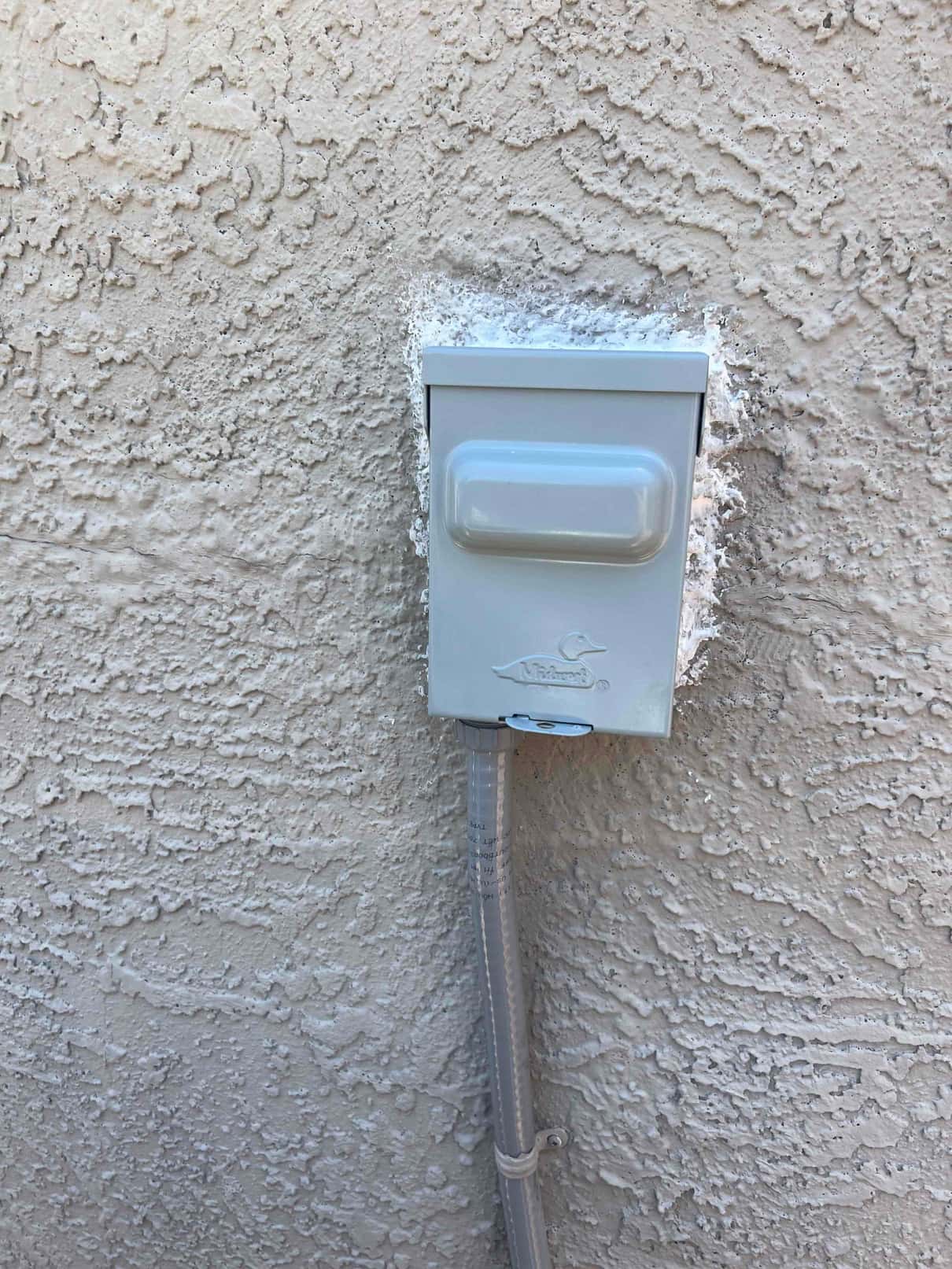 Weatherproof electrical disconnect box mounted on exterior stucco wall with sealed penetration and flexible conduit secured below.
