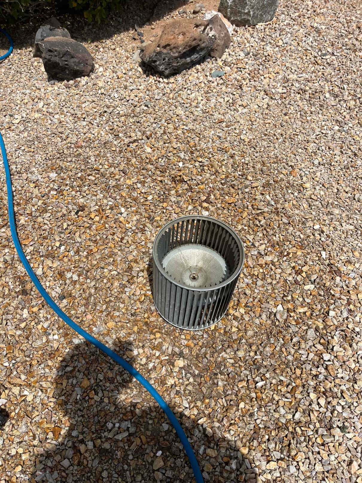 Blower wheel removed from HVAC system, sitting on gravel, showing dust buildup on blades and hub.